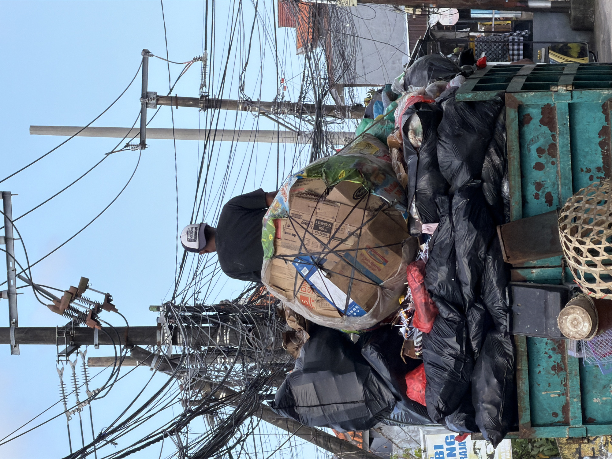 In Kuta Selatan, Indonesia, a truck is loaded with a large amount of trash, primarily black garbage bags and some cardboard boxes wrapped in plastic. A person wearing a black t-shirt and a white and black cap is visible from the back, standing on top of the load. The background features a chaotic arrangement of electrical wires and poles, indicative of a densely populated urban environment. Residential buildings with tiled roofs are also visible in the distance. The truck itself appears to be old, with visible rust on its teal-colored metal body. The sky is clear and blue.