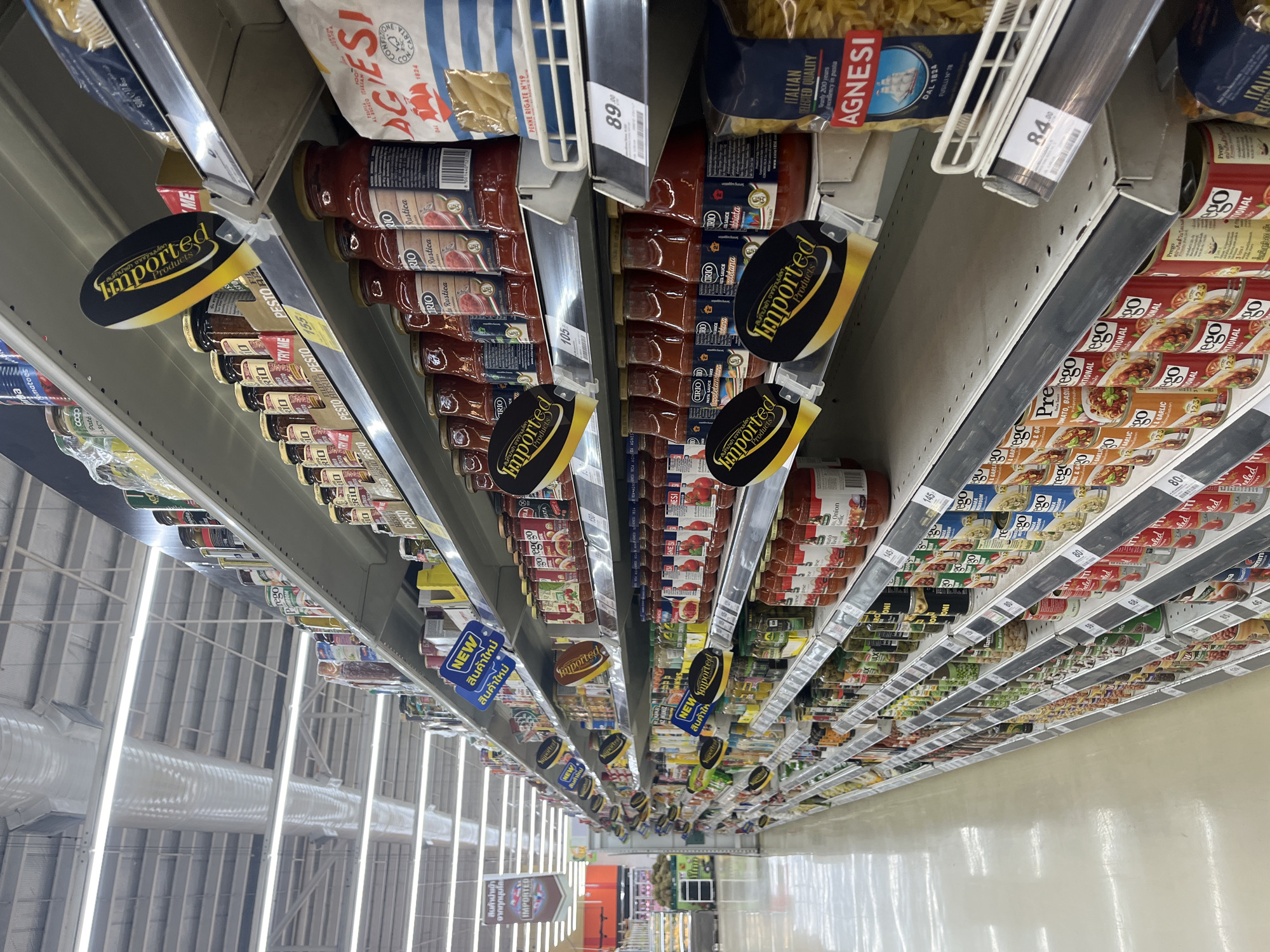 This is a low-angle, full shot of a well-lit aisle in a large supermarket in the city of Nong Pa Khrang, Thailand. The shelves are stocked with a variety of canned and bottled goods, primarily pasta sauces and canned food items. Many products are labeled as "Imported Products," indicated by oval-shaped yellow and black signs. The aisle extends into the distance, with bright fluorescent lights illuminating the scene from above. The floor is a light-colored, polished surface. No people are visible in the frame.