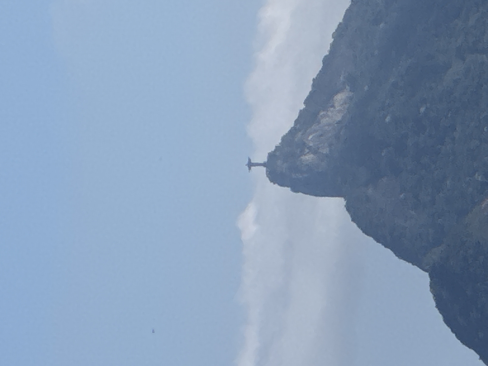 The image shows a distant view of the Christ the Redeemer statue atop Corcovado Mountain in Rio de Janeiro, Brazil. The statue, a large Art Deco sculpture of Jesus Christ, is silhouetted against a pale blue sky with scattered clouds. The mountain itself is covered in dark green vegetation. The statue is located in the city of Rio de Janeiro, not Niterói.