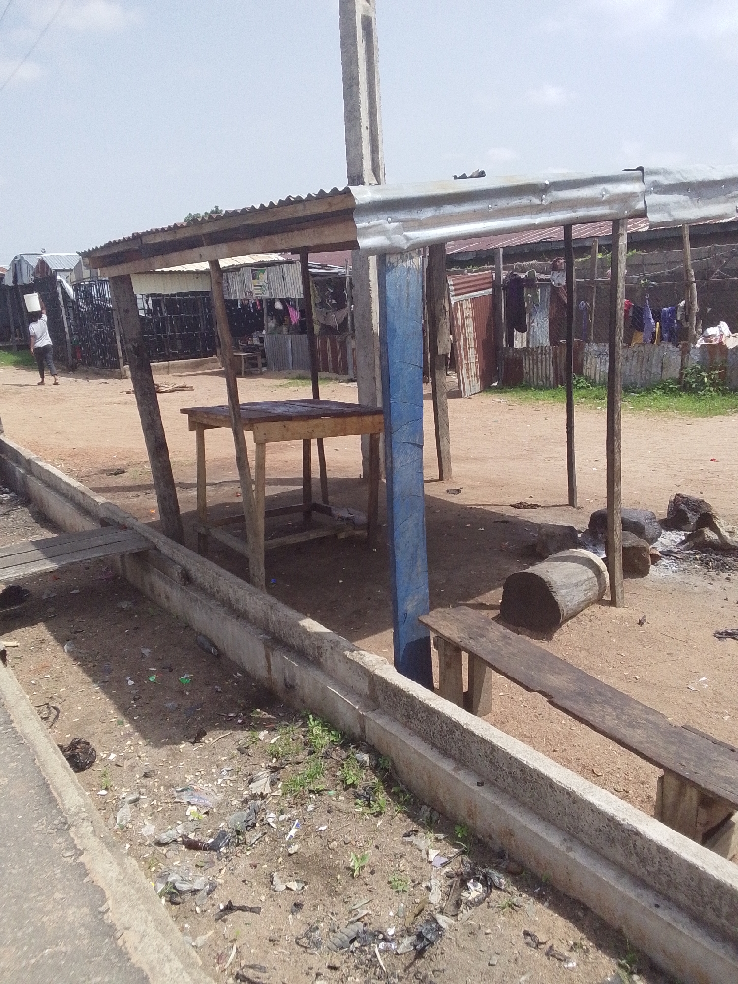 The image depicts an outdoor market stall area located in Jalingo, Nigeria. The scene is characterized by a makeshift shelter with a corrugated metal roof supported by wooden posts. Beneath the shelter, there is a wooden table with a rough-hewn top, suggesting a place for displaying goods or for customers. To the left, a person is seen walking away from the camera, carrying a white bucket on their head, a common sight in many African cities for transporting water or other items. The ground is sandy and dusty, littered with debris, including plastic waste and discarded materials. Adjacent to the shelter is a concrete drainage channel, also containing some litter. In the foreground, a long wooden bench is positioned, possibly for shoppers to rest. A large log, possibly used as seating or for a fire pit, is visible near the bench, alongside several rocks and remnants of a fire. In the background, more stalls or structures with corrugated metal walls are present, and laundry is hanging out to dry, indicating residential or commercial activity. The overall environment appears to be a somewhat unkempt but functional public space.