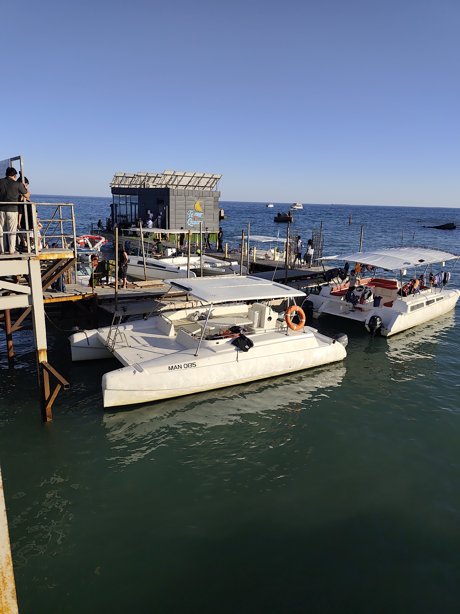 The image depicts a scenic view of a pier with several boats docked and people milling about. The scene appears to be set at a harbor or a beach resort, with clear blue skies and calm, greenish water. The time of day is likely late afternoon or early evening, as indicated by the warm, soft lighting and long shadows.
Several white catamarans are the most prominent objects in the foreground, with one bearing the visible text "MAN 0135" on its hull. These boats are equipped with canopies and appear ready for or have recently completed passenger transport. A small building with a modern design, featuring large windows and a sloped roof with solar panels, is situated on the pier. The building has a sign with a logo and the text "Te Quiero Cafe".
People are seen on the pier and on the boats, engaging in various activities such as boarding, disembarking, or socializing. Their presence suggests a lively atmosphere, possibly a tourist destination. The weather is pleasant and sunny, contributing to the overall relaxed and inviting ambiance of the scene. The setting is maritime, with the vast ocean stretching into the horizon.