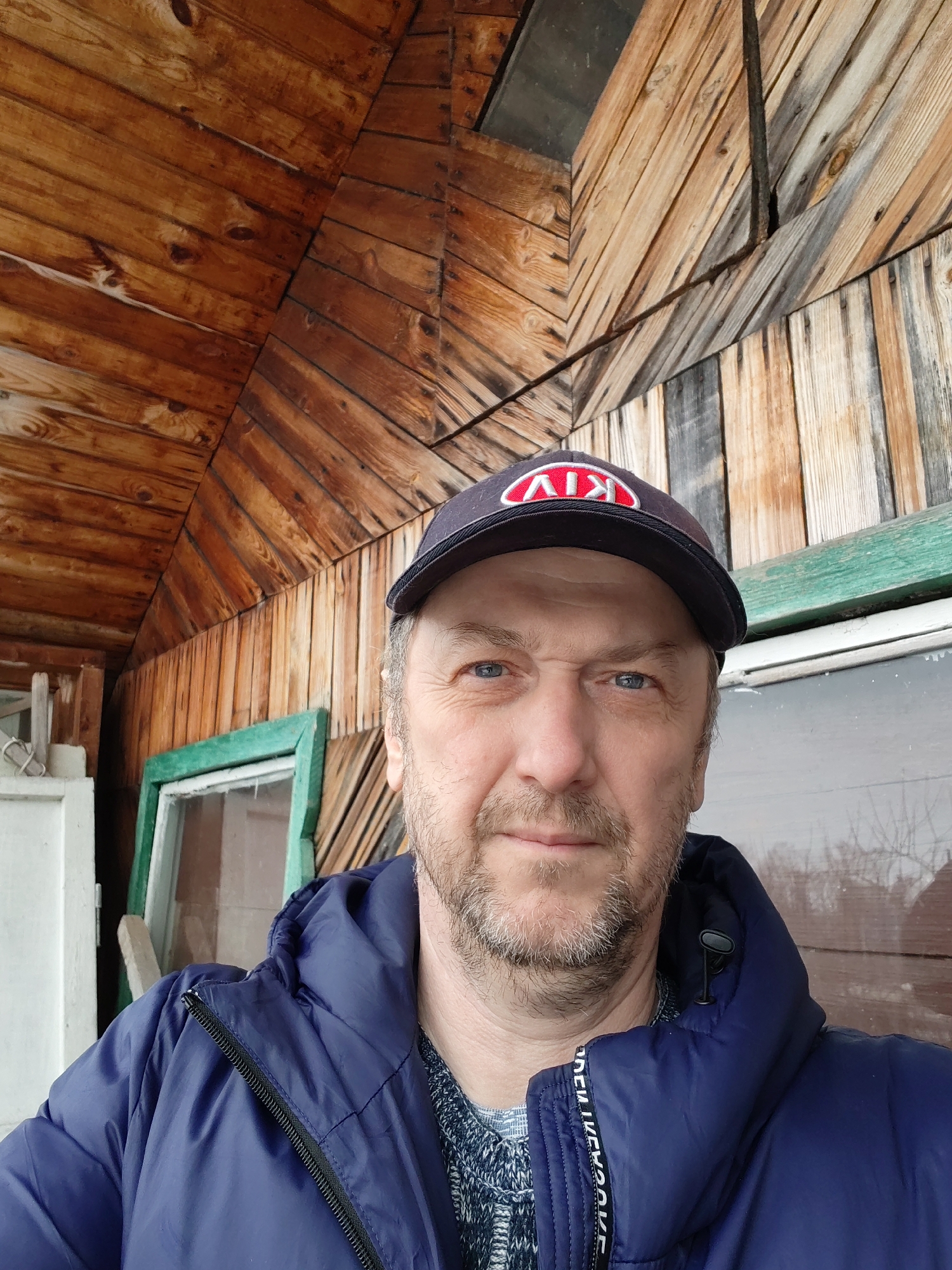 A middle-aged man is centered in the frame, wearing a blue puffy jacket and a black baseball cap with a red and white logo. He has short gray hair, a light beard, and blue eyes. The background consists of a wooden structure, likely an exterior wall and roof overhang of a building. The wood panels are arranged in a diagonal pattern and vary in color from light brown to gray. There is a window frame painted green visible on the left. The location is Chaikovsky, Russia.