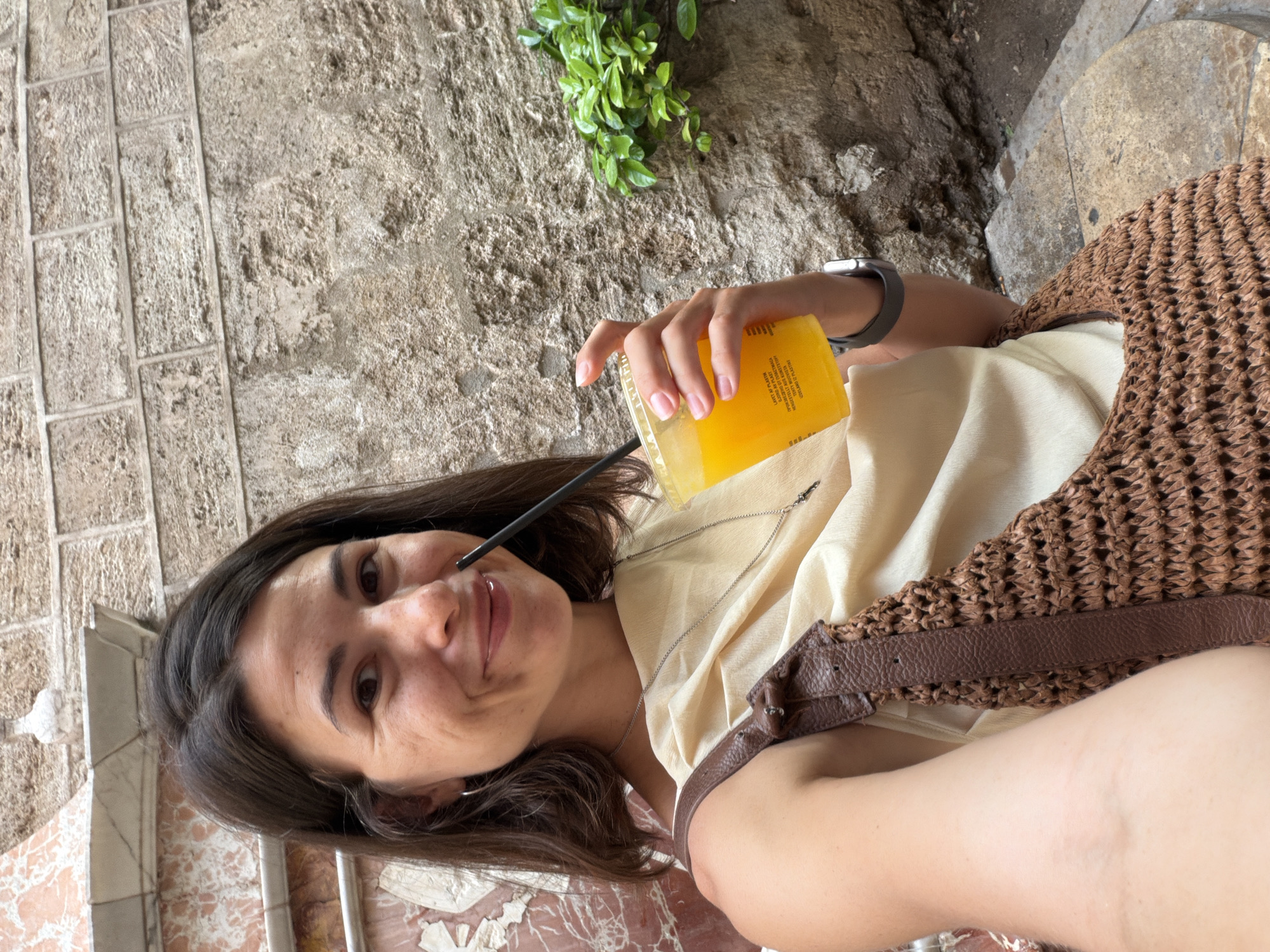 A woman with medium-length dark brown hair is taking a selfie while holding a plastic cup of orange juice with a black straw. She is smiling and looking at the camera. She is wearing a light beige, draped top and a brown woven bag with leather straps. A silver necklace with a small charm is visible. On her left wrist, she wears a smartwatch with a gray band. She is sitting outdoors in front of a textured, light-colored stone wall. To the right of the woman, a small cluster of green leaves is growing out of a crevice in the wall. The background also shows elements of marble and brickwork. The lighting suggests an overcast day or a shaded area.
