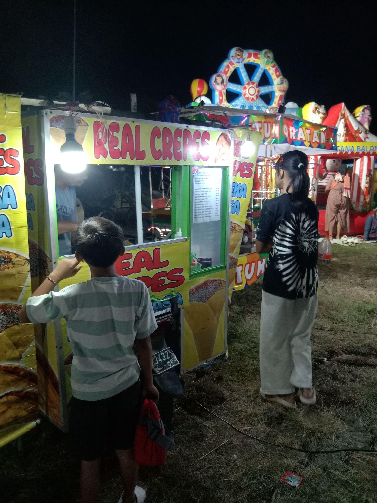 The image captures a nighttime scene at what appears to be a local fair or market in Stiaji, Indonesia. In the foreground, a young boy wearing a striped t-shirt and black shorts faces away from the camera, reaching towards a food stall. Next to him, a young woman with her hair in a ponytail, wearing a black t-shirt with a white spiral design and light-colored pants, stands looking towards the same stall.
The food stall, primarily yellow and green, displays prominent red lettering that reads "REAL CREPES." A menu is visible behind a glass partition. Another stall, further in the background, has "TUM" and "TANA BALON" written on it. Various colorful carnival attractions, including a Ferris wheel-like structure with painted faces and balloons, are visible in the background, suggesting a festive atmosphere. The ground is covered in dry grass and some scattered debris. The lighting is a mix of artificial lights from the stalls and attractions, illuminating the scene against the dark night sky. A motorcycle with a license plate reading "3432 05X" is parked next to the crepes stall.