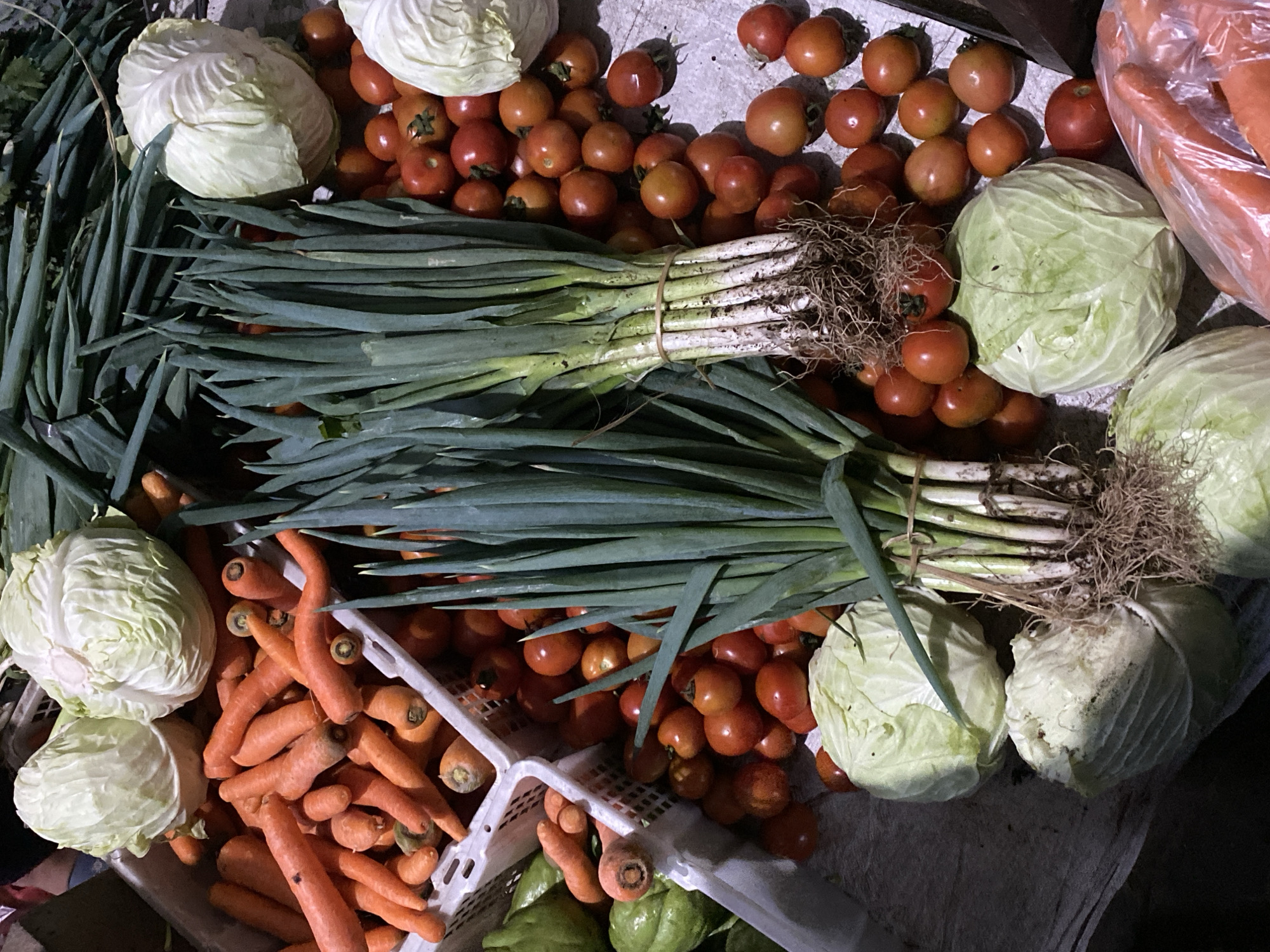 This is a close-up, overhead shot of a market stall in Ambarketawang, Indonesia, filled with fresh produce. The scene is dominated by piles of vegetables including several heads of white cabbage, bunches of long green onions, and numerous small red tomatoes. Carrots, some whole and some already separated, are visible in white plastic crates. A few green chayote squashes are also present. The produce is laid out on a grey surface, likely a table or ground cloth, with a hint of a woven basket in the background. The lighting suggests an outdoor market setting, possibly during daylight hours. There are no people or visible actions depicted in the frame.