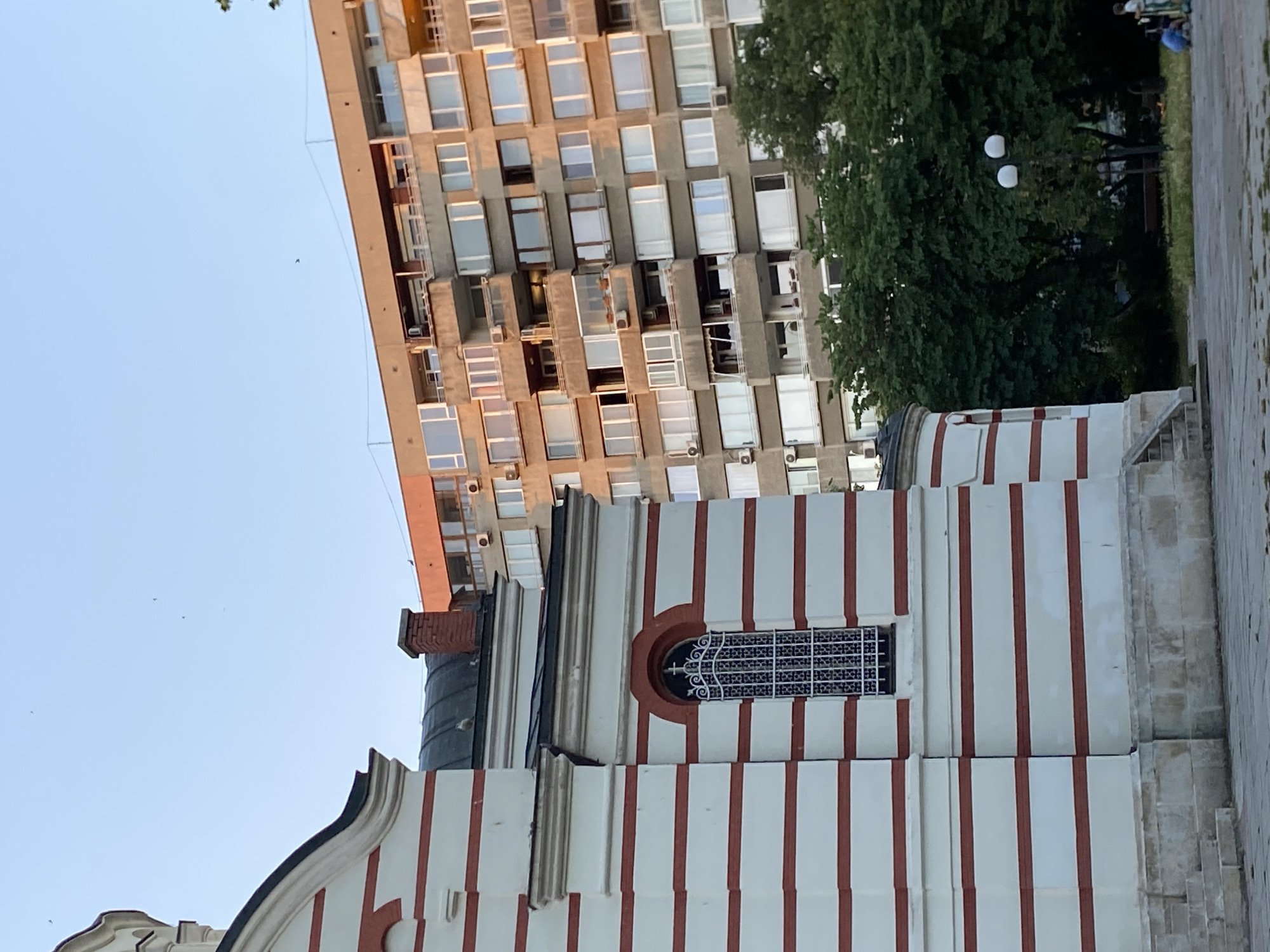 This media file is a photograph taken outdoors in Varna, Bulgaria. The image features a juxtaposition of old and new architecture. In the foreground, a building with white walls and red vertical stripes is visible. It appears to be a historical or religious structure, with a distinctive window featuring intricate metalwork. Behind this building, a modern, multi-story apartment complex with numerous windows dominates the upper portion of the frame. Trees and a lamppost with two spherical lights are also present, suggesting an urban park or a public space adjacent to the buildings. The sky is clear and blue, indicating it is daytime, likely late afternoon or early evening given the soft lighting. There are no people visible in the frame, and the overall atmosphere is serene and architectural. The image is oriented vertically, with the sky at the top and the ground at the bottom.