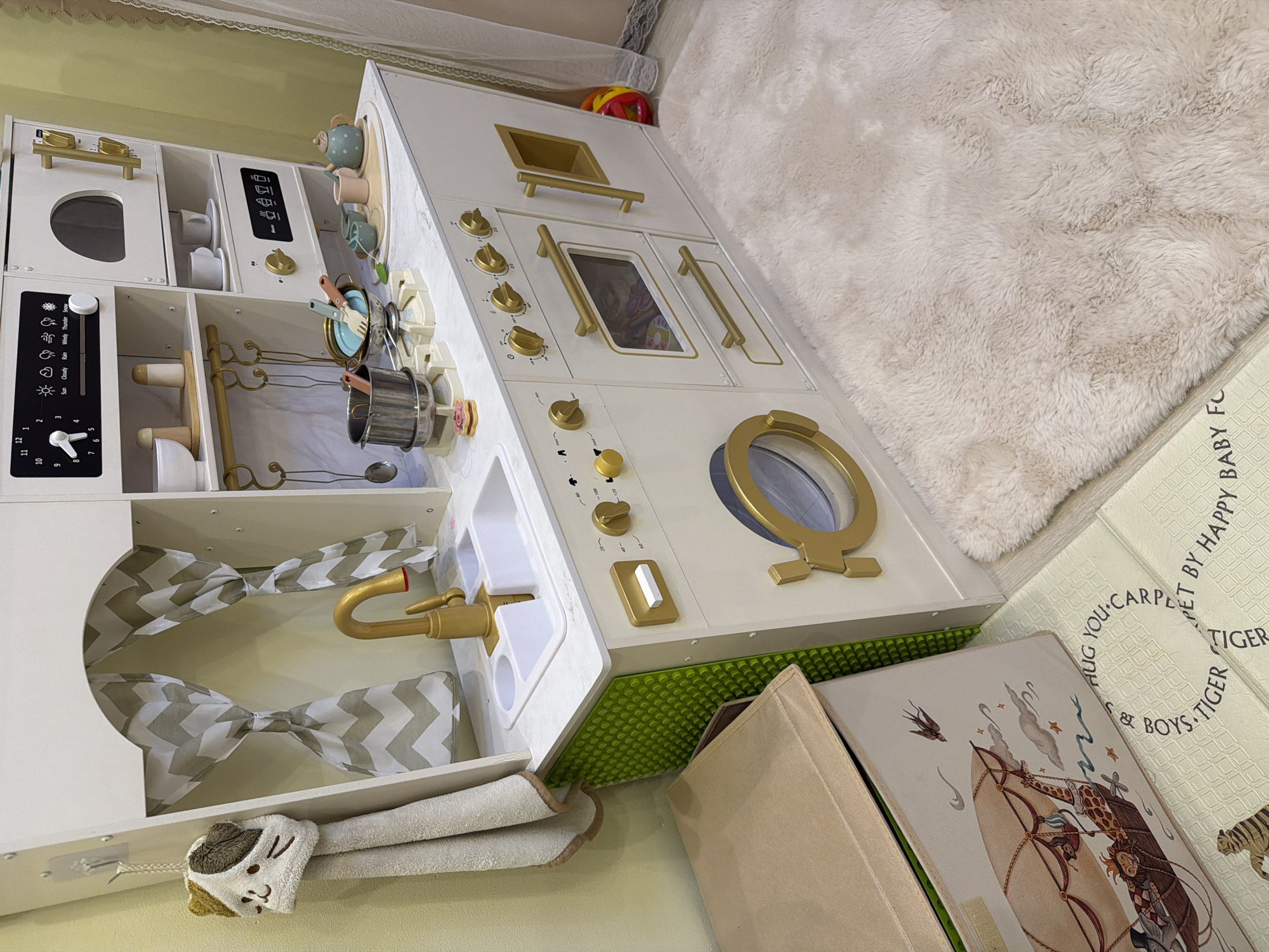 This media file features a child's wooden play kitchen, a popular toy for imaginative play. The kitchen is predominantly white with gold accents, suggesting a modern and elegant design. It includes various features mimicking a real kitchen, such as an oven, stovetop, sink with a gold faucet, and a control panel with dials and buttons. Several play accessories are visible, including pots, pans, utensils, cups, and a teapot. The play kitchen is set up on a light-colored, plush rug, possibly in a child's playroom or bedroom. The setting appears to be indoors, with a soft, diffused light, indicating it's likely daytime but not directly lit by the sun. There are no people visible in the image, focusing solely on the toy and its surroundings. In the background, a hint of a window with lace curtains can be seen. The overall impression is one of a well-equipped and inviting space for a child's pretend play. A few book covers with illustrations and text are also partially visible in the foreground. The text on one of the book covers reads "YOU CARPE S & BOYS TIGER" and "PET BY HAPPY BABY FO".