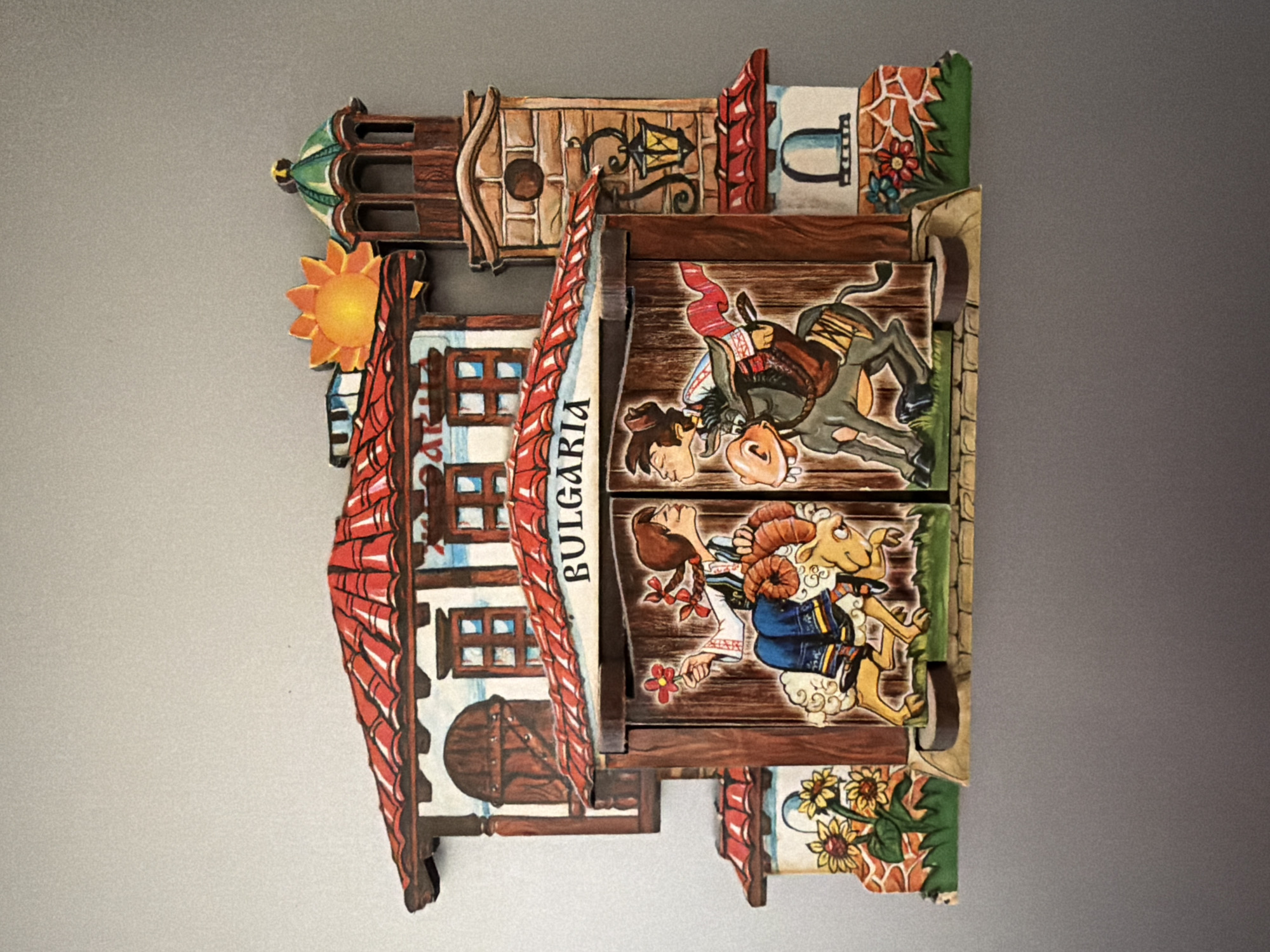 The image displays a colorful, three-dimensional decorative piece depicting a scene from Bulgarian culture. The central focus is a wooden facade with the word "BULGARIA" written across the top. On the left side of the facade, a woman wearing traditional Bulgarian clothing rides a sheep and holds a flower. On the right, a man on a donkey, also in traditional attire and holding a musical instrument, is depicted. Behind the facade, a building with red-tiled roofs, windows, and shutters is visible. To the right of the building is a brick tower with a green dome and a lantern. A sun is positioned in the upper right corner. The foreground features stylized sunflowers and other flowers. The piece is mounted against a plain gray background. No specific location in Pazardzhik, Bulgaria, can be identified from the image.