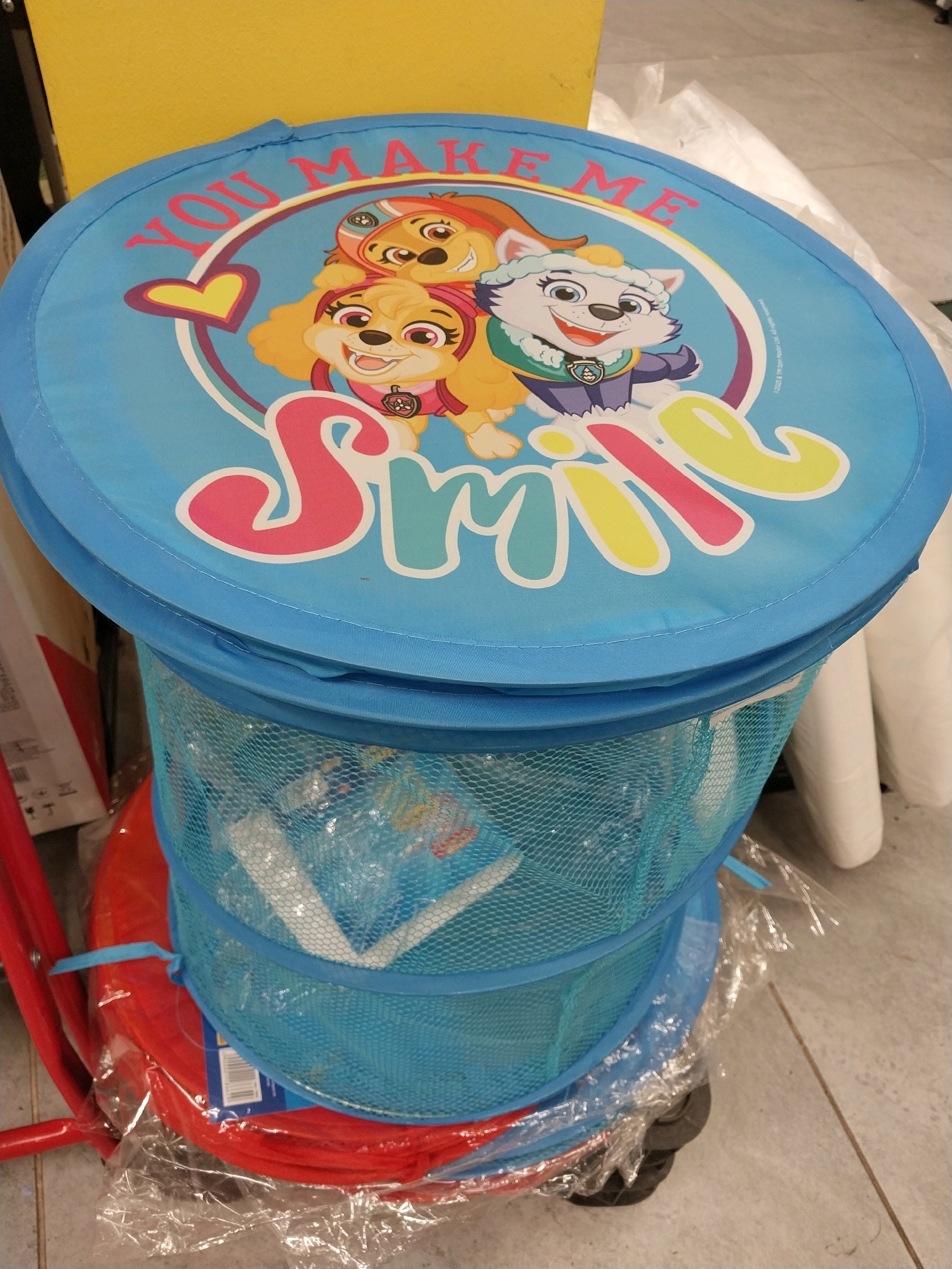 The image shows a blue pop-up laundry hamper with a Paw Patrol design. The design features three characters: Skye, Everest, and a third character who appears to be Ryder. The text "YOU MAKE ME SMILE" is prominently displayed on the lid of the hamper. The hamper is partially filled with white items wrapped in plastic. Below the hamper, a red circular object, possibly another hamper or a mat, is visible, also wrapped in plastic. The hamper is placed on a tiled floor, and a portion of a yellow wall is visible in the background. The scene appears to be indoors, possibly in a retail or storage area. The image was taken in the city of Dnipro, Ukraine.