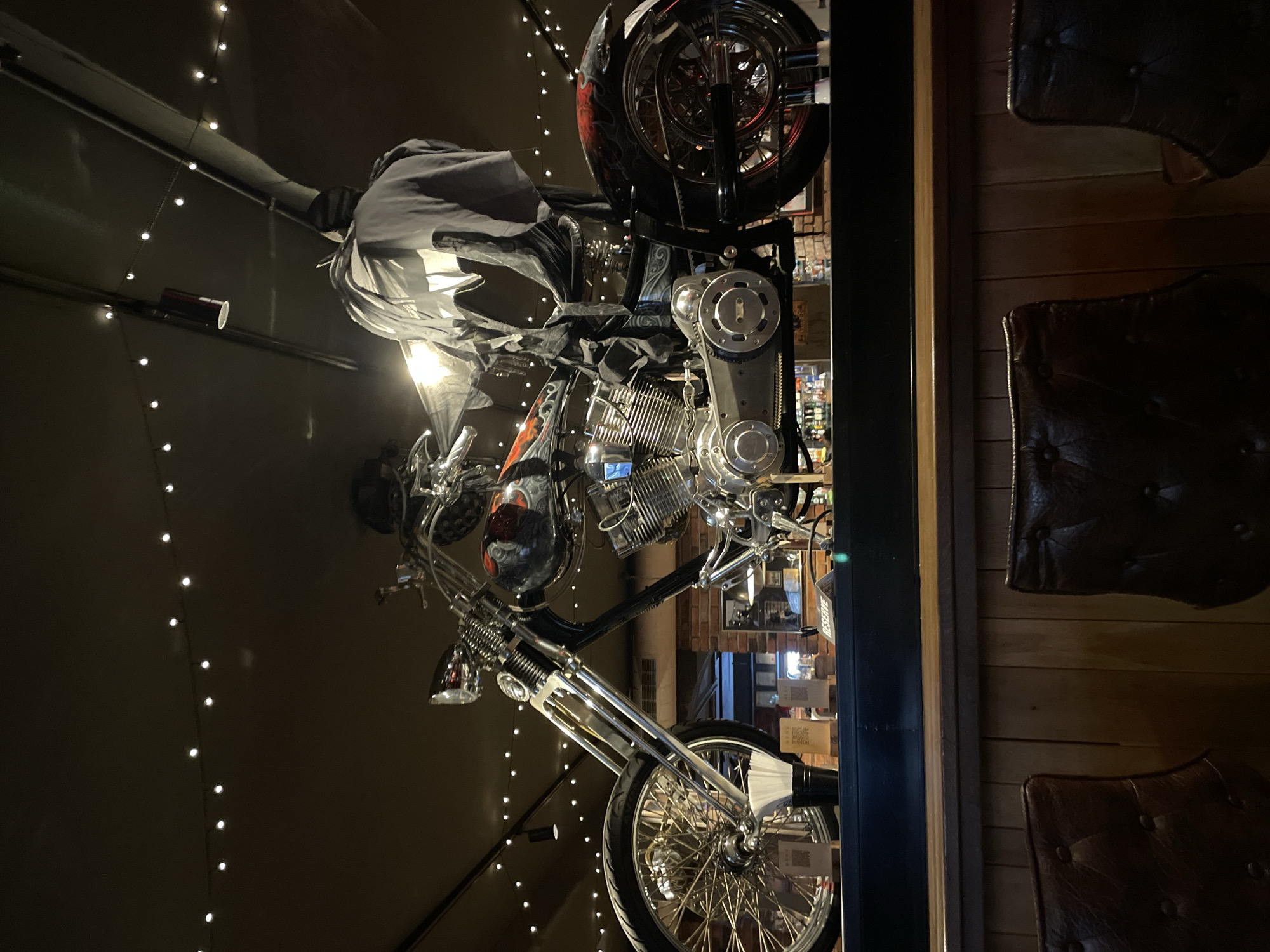 The image depicts a custom motorcycle mounted on a wall as decor, seemingly inside a bar or restaurant. The motorcycle features a chrome-finished engine and front forks, with black and red accents on the gas tank and rear fender. A dark cloth or cover is draped over the handlebars and front part of the motorcycle. The surrounding environment is dimly lit, illuminated by string lights that create a festive atmosphere. Next to the motorcycle, wooden panels and leather chairs can be seen, suggesting that the location is a pub or restaurant. It cannot be confirmed if the location is in Veliko Tarnovo, Bulgaria.