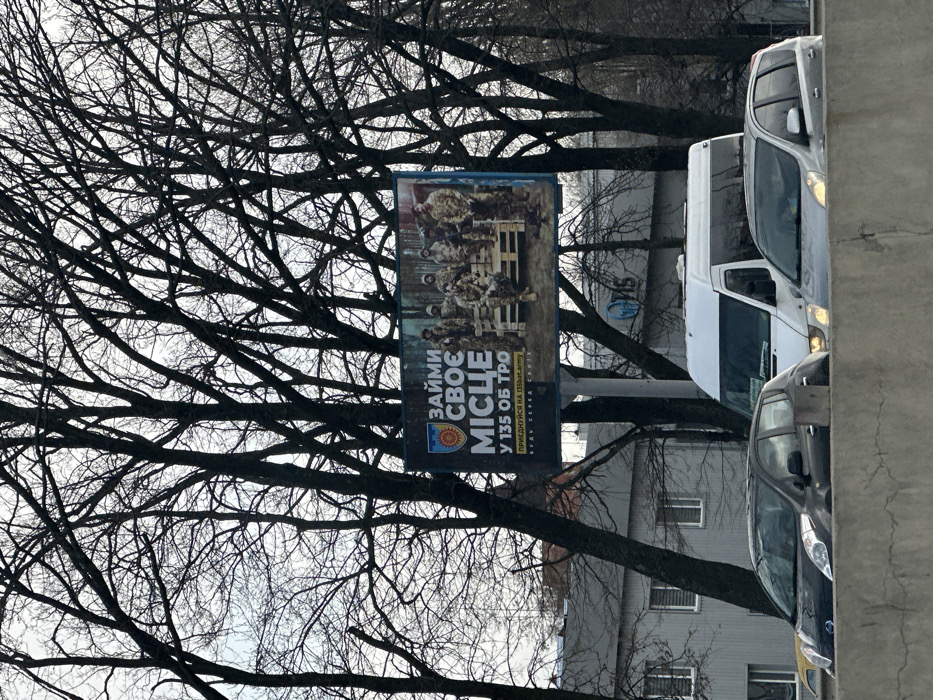 The image shows a billboard with a military recruitment advertisement. It features a group of soldiers wearing camouflage uniforms and holding rifles, standing on a pallet. The text on the billboard says "Займи своє місце у 135 ОБ ТРО" which translates to "Take your place in the 135th Territorial Defense Brigade". The billboard is located on a street in the town of Chabany, Ukraine, which is situated just southwest of Kyiv. Bare trees line the street in front of the billboard. A white van, an older model grey car, and a silver sedan are visible in the foreground. The image was likely taken sometime during the 2022 Russian invasion of Ukraine.