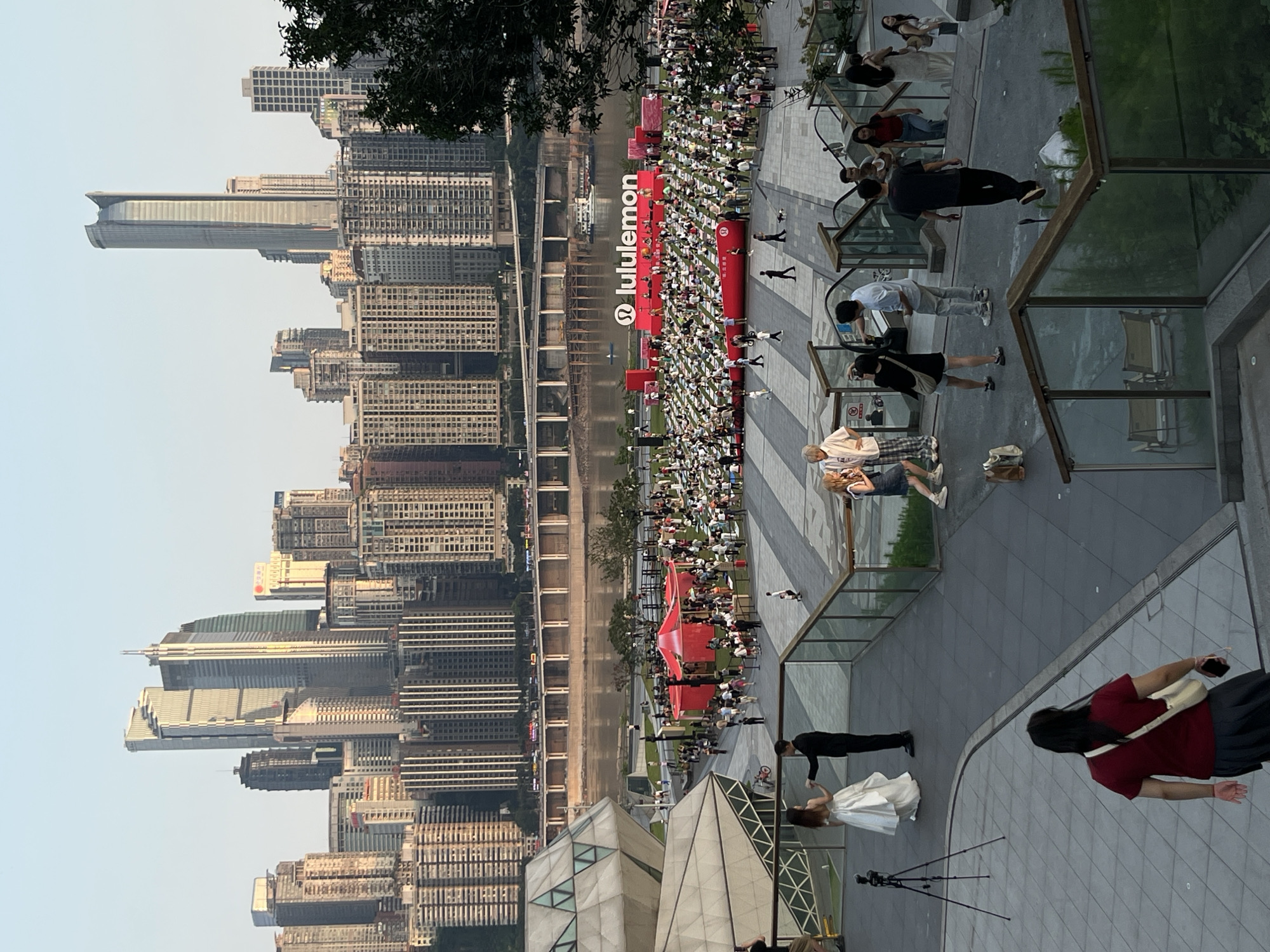 The image captures an outdoor event with a large group of people gathered in an open space in front of a city skyline. The setting appears to be a modern urban area, possibly a public plaza or park. In the background, numerous tall skyscrapers dominate the horizon, indicative of a major city. A wide river flows in front of the buildings, crossed by a bridge.
In the foreground and midground, a large number of people are seated on the ground, seemingly participating in an organized activity, possibly a yoga or meditation session, as suggested by the presence of mats and a large "lululemon" banner on a stage. The stage is decorated with red fabric and has a platform where performers or speakers are visible. Red tents are also set up around the event area.
Several individuals are in motion on the pathways and stairs surrounding the gathering. A couple dressed formally, a woman in a white gown and a man in a black suit, are positioned on a raised walkway, possibly posing for a photograph, with a tripod and camera visible nearby. Other individuals are walking, talking, and observing the scene. The architecture of the buildings in the foreground is modern and geometric, with glass facades. The overall atmosphere suggests a well-attended public event in a vibrant, contemporary cityscape. The specific location, 江北城街道, China, is a district within Chongqing known for its modern architecture and riverside developments.