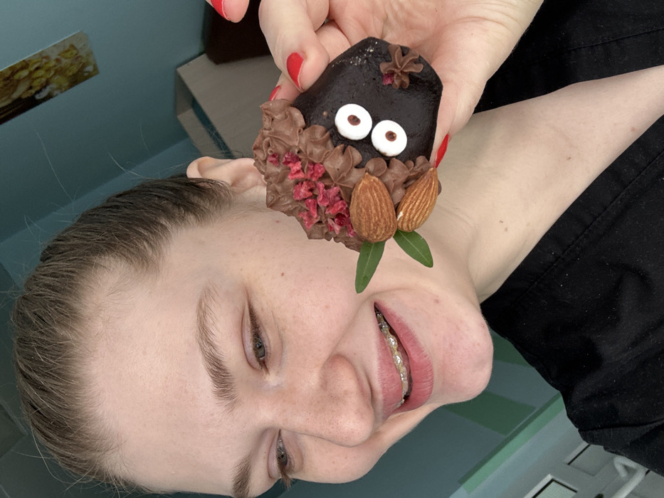 A medium close-up image depicts a person holding a decorated dessert in what appears to be an indoor setting in Kazan, Russia. The individual's head is tilted back and to the right, featuring fair skin, light brown hair, and a visible smile or laugh that reveals dental braces on the upper teeth. Their lips are tinted pink. A hand, adorned with red nail polish, holds a dark brown, irregularly shaped dessert item. This dessert is decorated with two white circular sugar eyes with black pupils, brown piped frosting, multiple whole almonds, small green leaf garnishes, and reddish-pink granular toppings. The person is wearing a dark-colored garment, partially visible on their upper torso. The background consists of a solid light teal-colored wall, with a segment of a rectangular framed artwork, exhibiting yellow and gold tones, partially visible in the lower-left corner of the frame.
