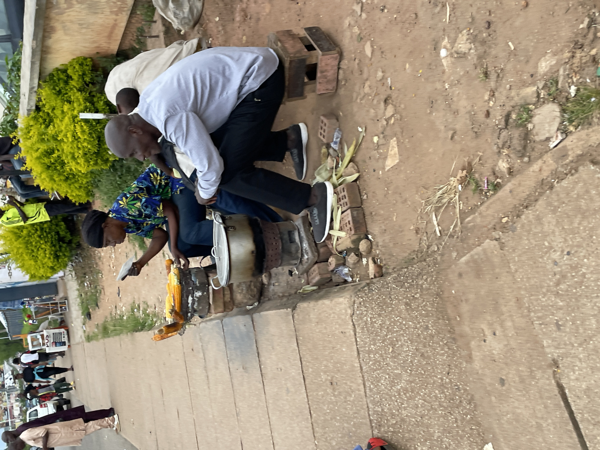 The image depicts a street scene in Jos, Nigeria, likely in a busy marketplace or roadside area. In the foreground, two individuals are seated. The woman on the left, wearing a headscarf and a colorful patterned shirt, is holding a small item, possibly a menu or money, and looking towards the right. In front of her is a makeshift cooking setup with corn cobs roasting on an open fire. A large metal pot sits on the side. To her right, a man in a light-colored shirt and dark pants is seated on a small wooden stool, looking to his left. He is wearing black sneakers. Behind them, more people and a white structure resembling a food stall are visible. The ground is a mix of dirt and concrete, with scattered debris and vegetation. The overall atmosphere suggests a typical urban environment with informal commerce.