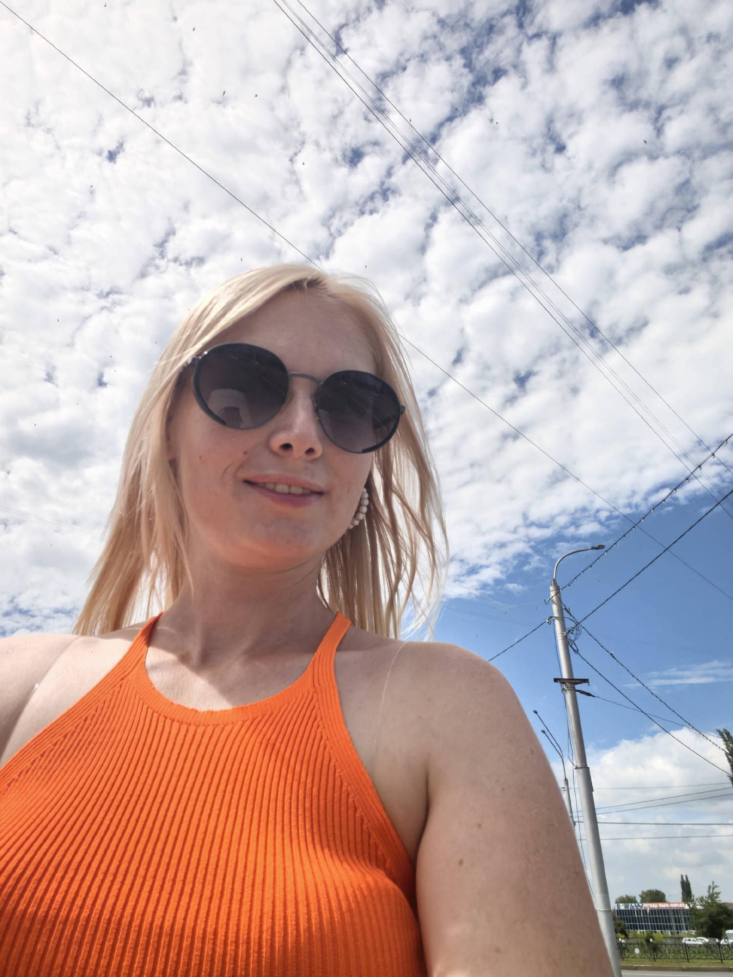 The image is a selfie taken outdoors, likely on a street in Sterlitamak, Russia, given the context of the request. A blonde woman wearing large, dark-rimmed sunglasses and an orange ribbed halter top is the primary subject. She has a faint smile on her lips and is wearing pearl earrings. Her hair is blonde and falls to her shoulders. The background consists of a bright blue sky filled with fluffy white clouds. Overhead, multiple power lines are visible, crossing diagonally across the frame. To the right, a street light pole with attached wires stands prominently. In the distance, a building with a blue facade and some greenery can be seen, suggesting an urban or suburban environment. The lighting indicates it is daytime, with the sun casting shadows and highlighting the woman's features.