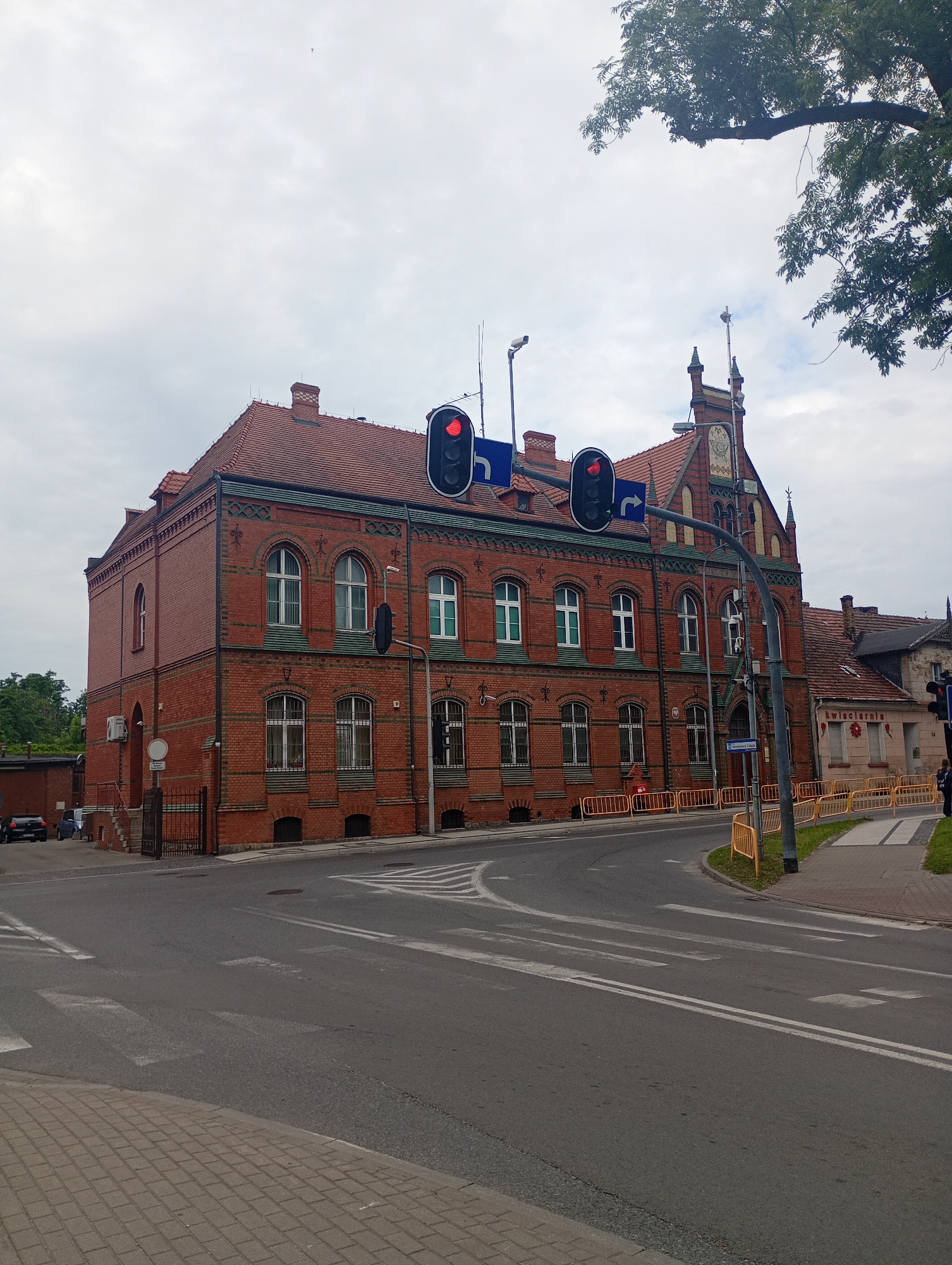 The image shows a red brick building in Międzyrzecz, Poland, likely the former Post Office building, located at Plac Jana Pawła II. The building has a red tile roof, arched windows with white frames, and decorative green and black brickwork. A traffic light is visible, showing a red signal. In front of the building, there are yellow safety barriers and a pedestrian crossing marked on the asphalt road. The sky is overcast. To the right of the main building, a smaller building with a sign that reads "kwiaciarnia" (florist) is visible. A tree with green leaves partially obscures the top right corner of the image. There are no people clearly visible in the foreground.