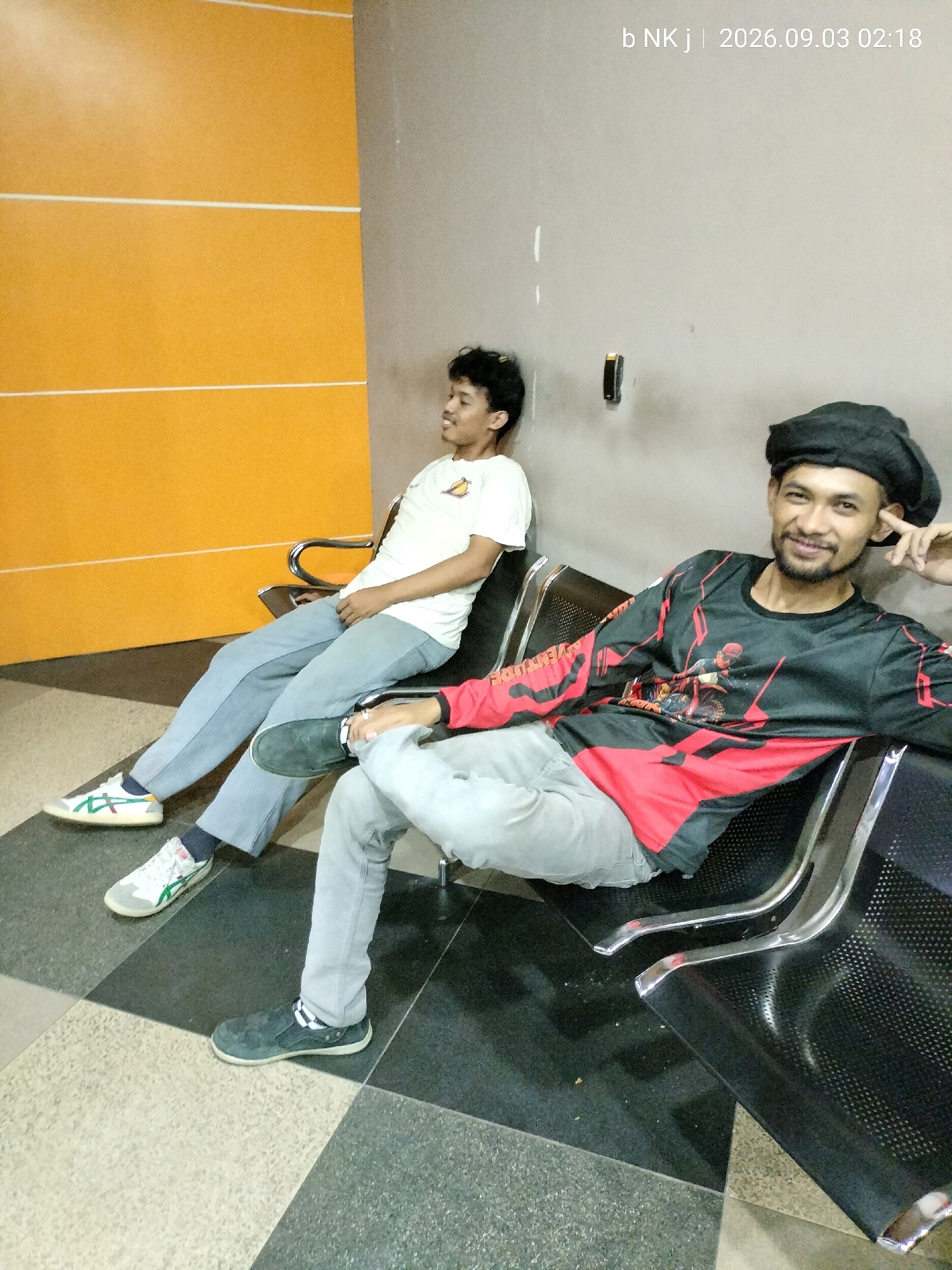 Two men are seated in what appears to be a waiting area. The man on the right is wearing a black beanie and a long-sleeved black shirt with red accents and graphics. He is smiling and looking towards the camera, with his right hand resting on his head. The man on the left is wearing a white t-shirt and light blue pants, looking away from the camera. Both men are seated on black, metal-frame chairs. The background features a textured orange wall with white horizontal lines and a plain gray wall. The floor is a checkered pattern of light and dark tiles. The timestamp "b NK j | 2026.09.03 02:18" is visible in the upper right corner, suggesting the image was taken in South Jakarta, Indonesia.