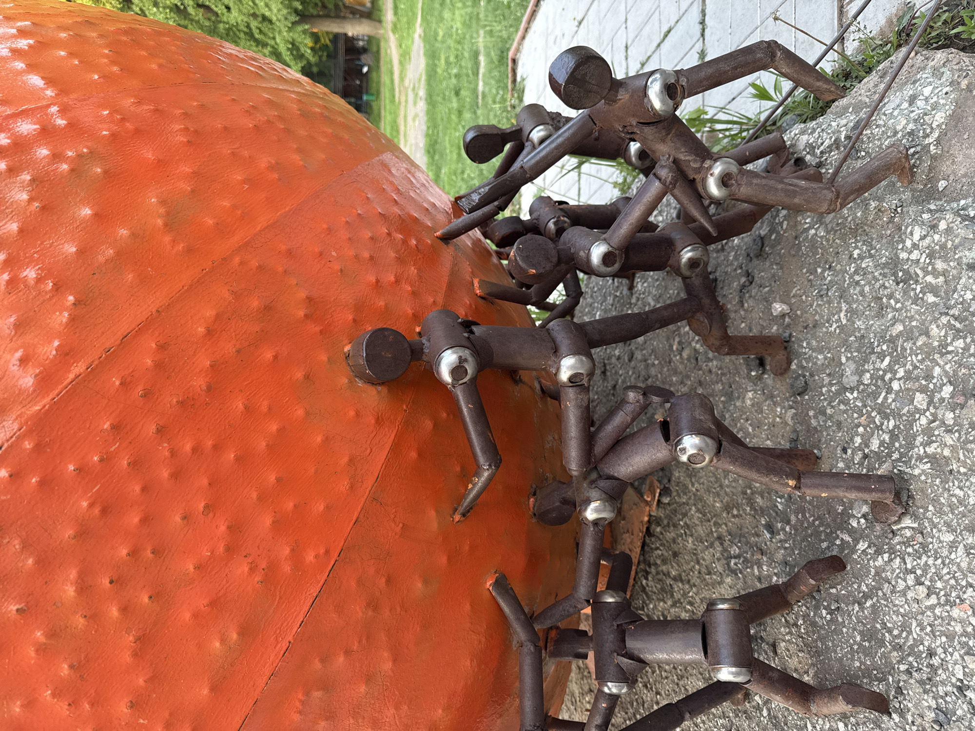 The image shows a close-up of an outdoor metal sculpture located in Izhevsk, Russia. The sculpture features a large, orange, textured sphere with numerous small, metal figures seemingly climbing or clinging to its surface. The figures appear to be made of dark, possibly rusted, metal and have a crude, stylized human form. The base of the sculpture rests on a weathered concrete surface. In the background, blurred greenery suggests a park or outdoor setting.
