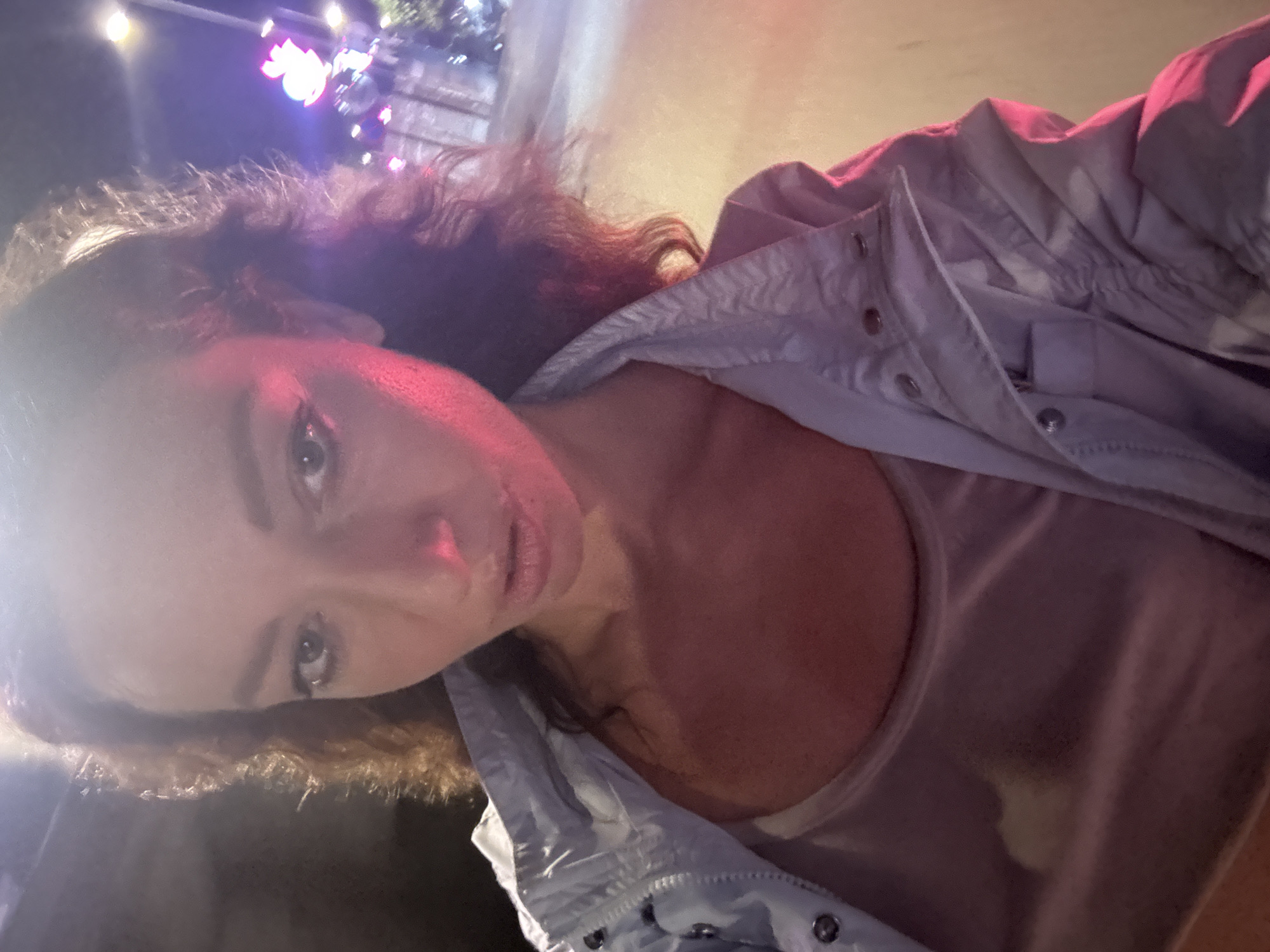 This is a selfie taken by a woman at night in Hanoi, Vietnam. She is wearing a light-colored, possibly light blue or grey, jacket over a sheer, light-colored top. The woman has dark, wavy hair and is looking towards the camera. Her face is illuminated by a strong pinkish-red light, likely from a nearby light source, which casts shadows and highlights on her skin. The background is dark, with blurred colorful lights visible, suggesting an outdoor setting possibly at a festival or entertainment area. One of the lights appears to be a bright, colorful ornament or decoration. The overall mood is candid and possibly energetic, given the lighting and selfie style. The time of day is clearly night.