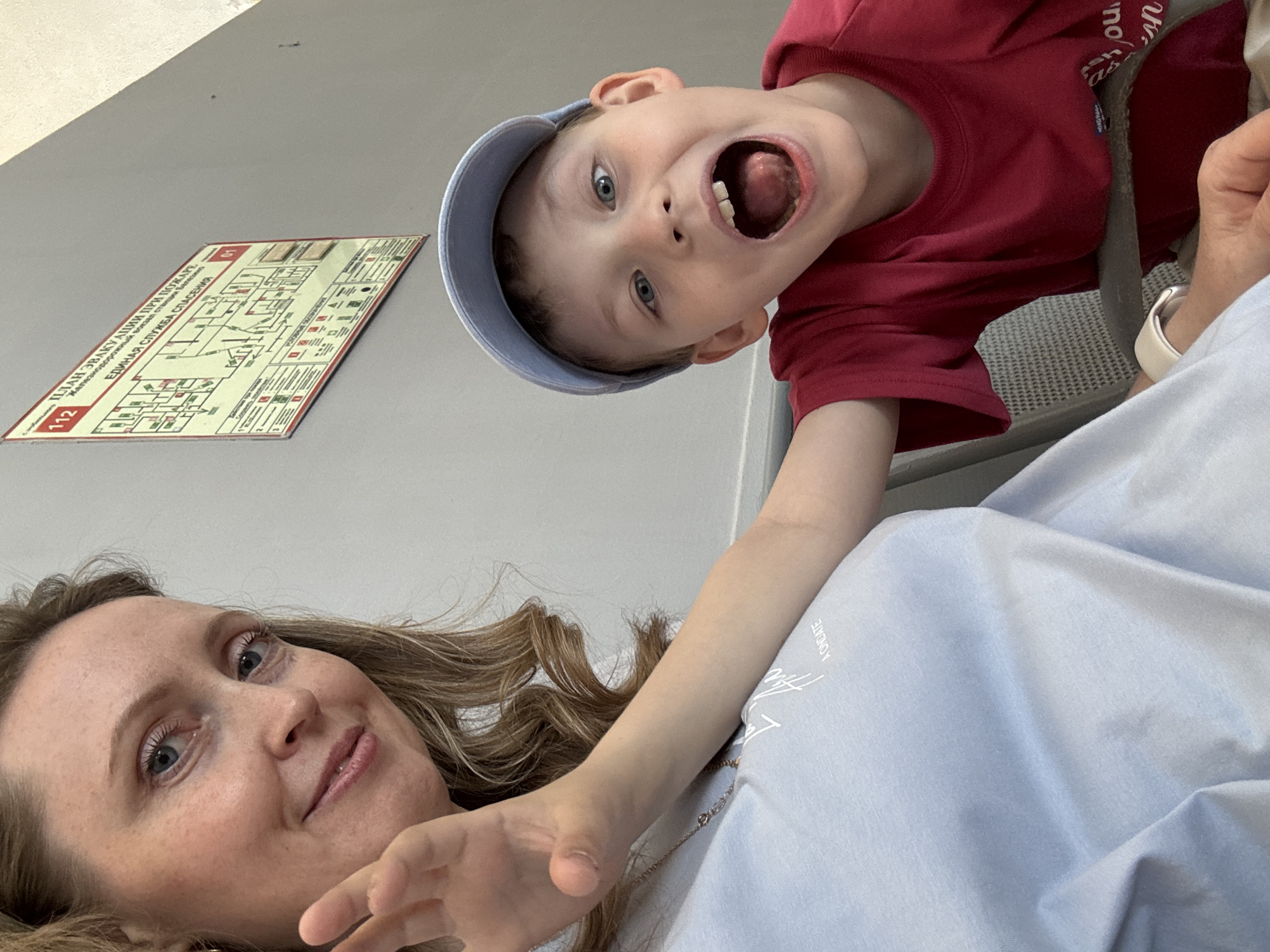 A woman and a young child are posing for a selfie. The woman is lying down, looking at the camera with a slight smile. The child is positioned above her, also looking at the camera with his mouth wide open. He is wearing a red shirt and a blue cap.
Behind them is a wall with a fire evacuation plan printed on it. The plan includes the text "ПЛАН ЭВАКУАЦИИ ПРИ ПОЖАРЕ" and "ЕДИНАЯ СЛУЖБА СПАСЕНИЯ," indicating that the image was taken in Russia. The text on the evacuation plan also includes "Железнодорожный вокзал станция лесовинь," which can be translated to "Railway station station forest," and "112" indicating it is a Russian railway station. The station is most likely in the city of Balezino, Russia, given the other text on the sign.