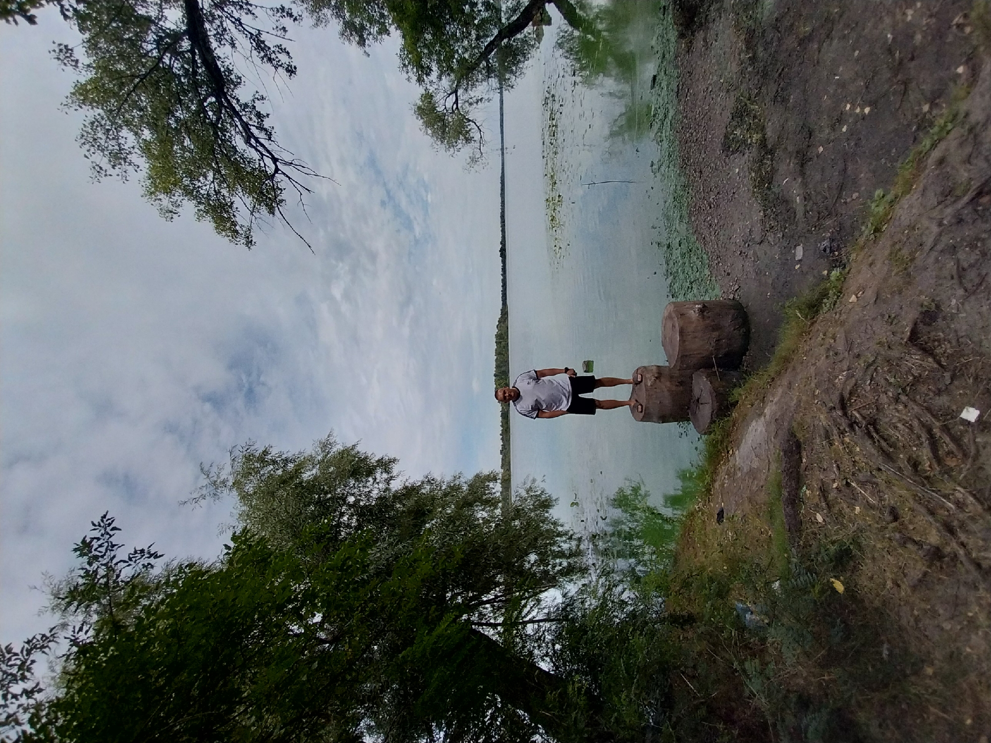 A full-body, wide-angle shot captures a man standing on two tree stumps on the bank of a body of water. The man, who appears to be in his 40s or 50s, is wearing a white t-shirt with a black logo and black athletic shorts. He is facing the camera with his hands in his pockets and a slight smile. The tree stumps are weathered and have a rough texture.
The body of water, likely a river or lake, is calm and reflects the overcast sky. Green aquatic plants dot the surface of the water, and further in the distance, a line of trees marks the opposite shore. The foreground shows a muddy bank with exposed tree roots and some scattered debris, including pieces of plastic. Lush green foliage, including large trees with dense leaves, frames the scene on both the left and right sides of the image, with branches extending over the water. The overall impression is a natural, somewhat untamed environment.
The image was taken in Dnipro, Ukraine. The specific location is on the bank of the Dnipro River, likely in a less developed, natural area, possibly a park or a less maintained recreational zone within the city. The presence of the river and the surrounding vegetation suggests a natural landscape within the urban environment of Dnipro.
