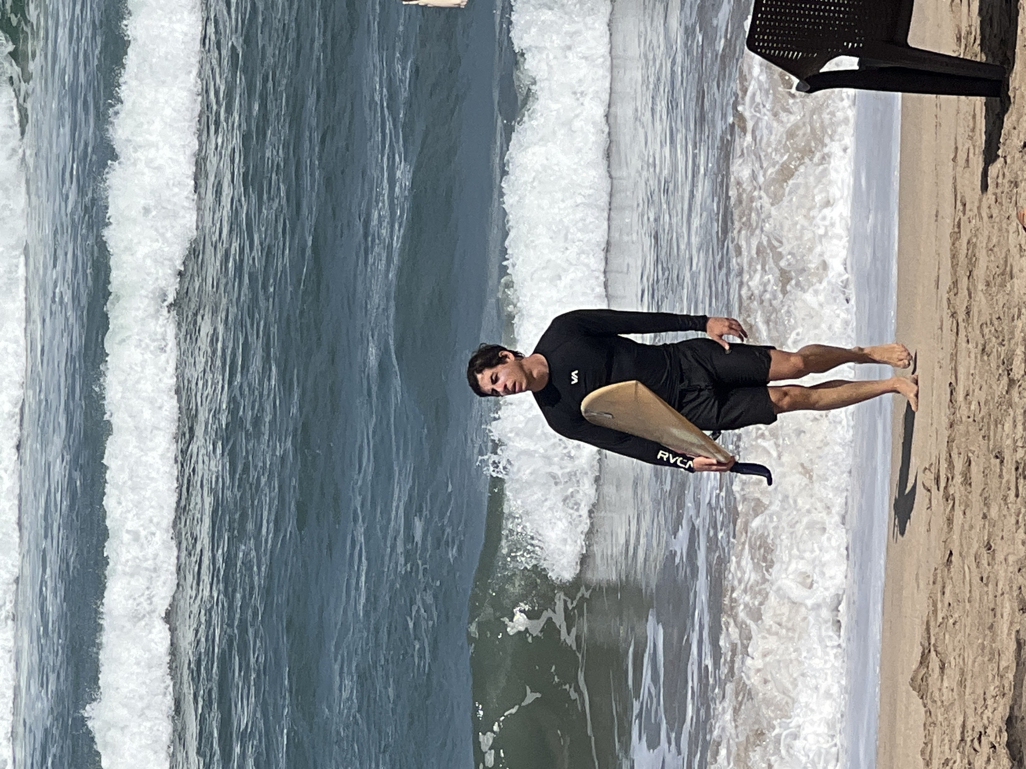A young man with dark hair, wearing a black long-sleeved rash guard with a white "VA" logo and black board shorts, walks along the wet sand at the edge of the ocean. He is carrying a light-colored surfboard under his right arm. The ocean is blue with white-capped waves breaking near the shore. The sand is light brown and shows some texture. In the background, there are multiple lines of waves. To the right of the frame, a portion of a dark plastic beach chair is visible. The overall scene depicts a surfer at Kuta Beach, Indonesia.