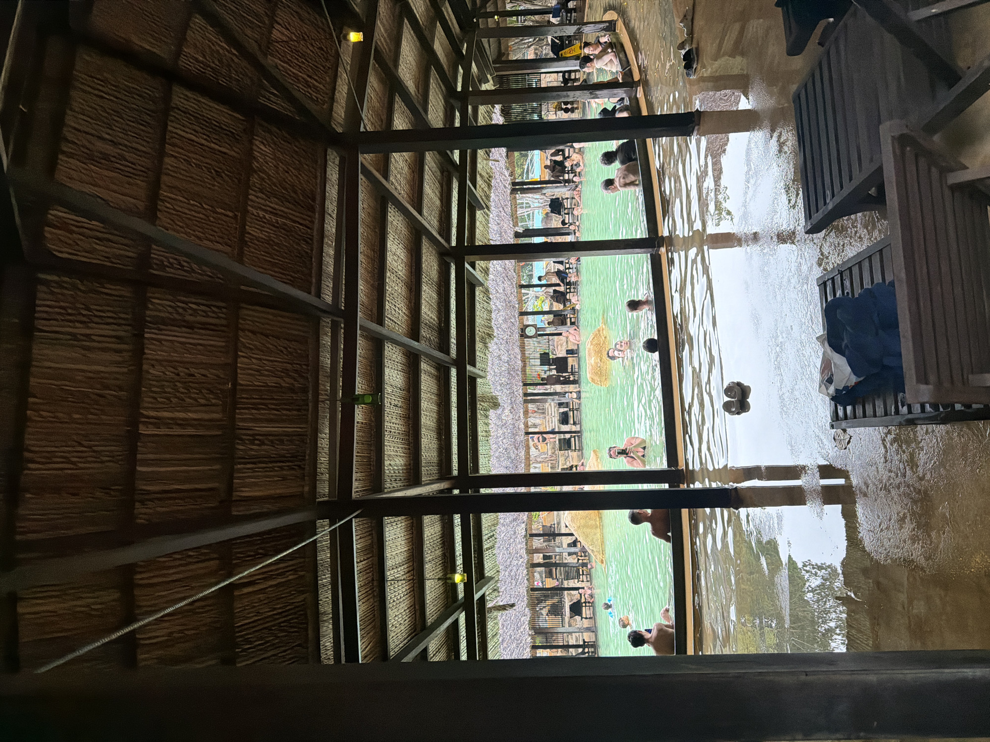 This is a medium shot, indoor photograph taken from a slightly elevated viewpoint looking into a geothermal hot spring pool area at a resort. The roof structure above is made of thatched panels supported by dark wooden beams and pillars. Natural light filters through the open sides of the structure.
In the foreground, there are two dark wooden lounge chairs with towels draped over them. The floor is wet and reflective, with visible puddles and water stains.
The main focus of the image is the large, shallow, turquoise-colored hot spring pool. Several people are seen bathing and relaxing in the water, with their torsos and heads visible. Some are interacting, while others are solitary. The pool is surrounded by a low barrier, beyond which are more thatched structures, lounge chairs, and large rocks.
The scene suggests a leisurely and relaxing atmosphere, typical of a spa or resort. Based on the architecture and ambiance, this appears to be the Suoi Khoang Nong hot spring at the I-Resort in Nha Trang, Vietnam, known for its geothermal pools and spa treatments.