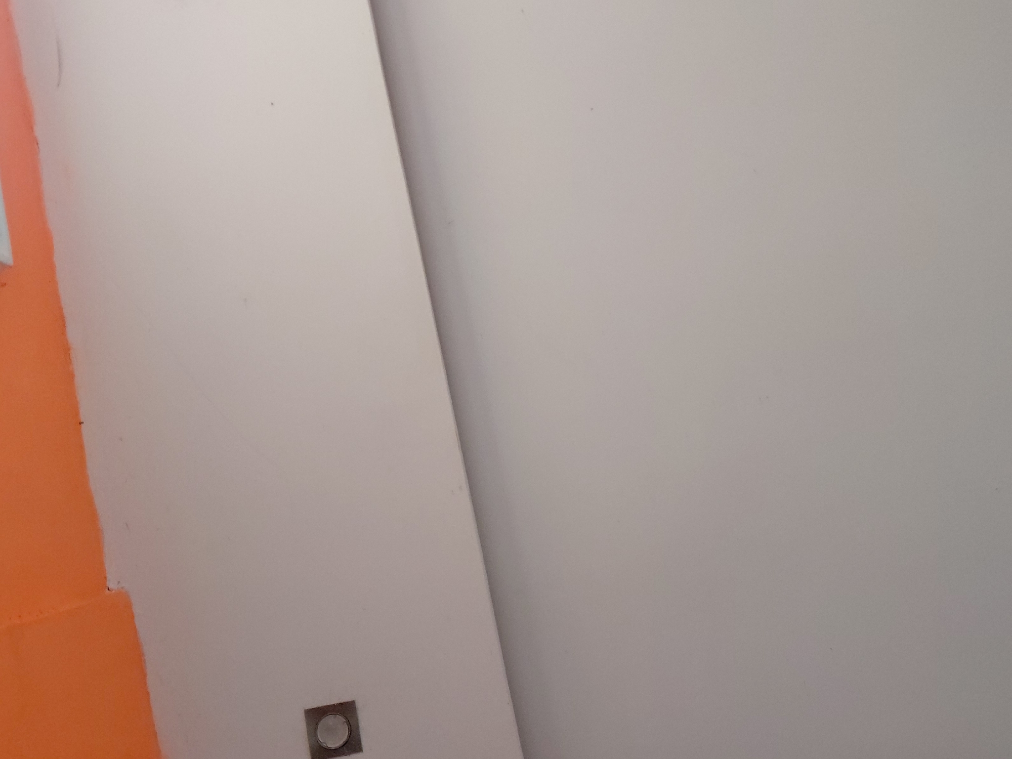 The image presents an abstract view of an interior space, primarily composed of plain architectural surfaces. On the left, a wall features a section painted in a vibrant orange, abruptly transitioning into a light grey or off-white area with an uneven paint line. This light grey surface forms a distinct, sharp corner or angled joint with another larger, uniformly light grey wall or panel that occupies the majority of the right side of the frame. The angle runs diagonally across the image, creating a sense of depth and separation between the planes.
Near the bottom-left of the central light grey surface, a small, square metallic fixture is embedded. It possesses a reflective, possibly brushed chrome or stainless steel, finish and contains a circular, light-colored or translucent component in its center, resembling a recessed light or a sensor. The surfaces are generally smooth with minimal, subtle imperfections. The lighting is even, suggesting an indoor environment. There are no people, objects beyond the described fixture, or activities depicted. No text is visible in the image. The location context is Santa Ana, Venezuela.