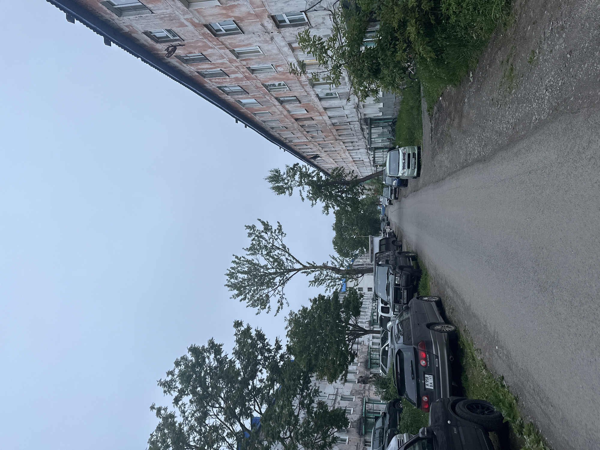 The image shows a street scene in Vilyuchinsk, Russia. On the right side of the image is a long, multi-story apartment building with a weathered, peeling facade, predominantly in shades of off-white and reddish-orange. The building has numerous windows, many of which appear to have white frames. The roofline is visible, with a dark, sloped covering and visible edges. Trees with green foliage grow along the front of the building and in front of the apartment complex on the left.
The street in the foreground is paved with asphalt and gravel, with a clear demarcation between the road surface and the grassy embankment. Several vehicles are parked along the right side of the street, adjacent to the apartment building, and along the left side, closer to the other apartment complex. These vehicles include SUVs and station wagons, some with visible license plates. Green grass and bushes are present along the edges of the street and in front of the buildings. The sky is overcast with a pale blue-gray hue. The overall impression is that of a residential area in a possibly older or less maintained part of the city.