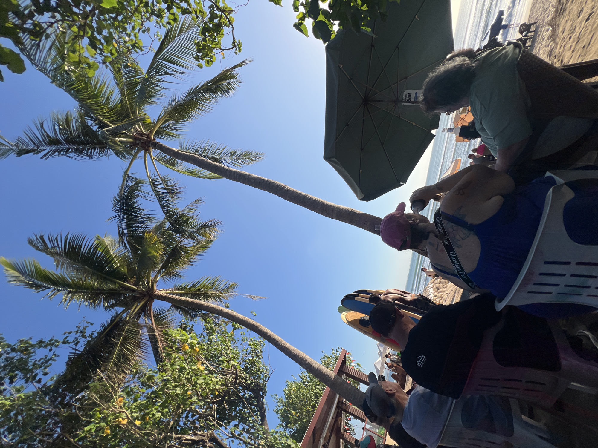 The image depicts a beach scene in Kuta, Indonesia, likely a cafe or restaurant area near the shore. In the foreground, several people are seated at tables on the sand, facing the ocean. Two tall palm trees frame the upper portion of the image, with their fronds extending into a clear blue sky. Some of the people are drinking from bottles. Surfboards are visible leaning against a structure to the left, suggesting proximity to a surf school or rental. An umbrella provides shade over some of the seating. In the background, the ocean is visible with gentle waves. The overall atmosphere suggests a relaxed, tropical setting.