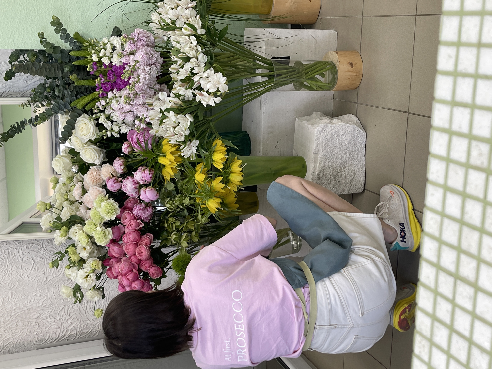 A high-angle shot displays a woman arranging flowers, seemingly indoors. The scene is in Izhevsk, Russia. The woman, wearing a pink t-shirt with "At first, PROSECCO" printed on the back, is kneeling beside multiple vases filled with various types of flowers. She is wearing blue coverings over her lower legs, white shorts, and Hoka running shoes. The flowers include roses, peonies, alstroemeria, and other types in shades of white, pink, purple, and yellow. The vases are sitting on stone blocks on a tile floor. The background is a light green wall. A window is visible in the background.