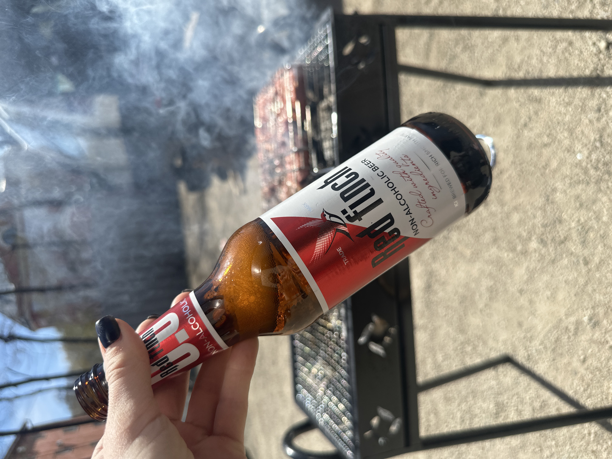 A hand with dark nail polish holds a bottle of "Red Finch" non-alcoholic beer against a background with smoke and a barbecue grill. The scene appears to be outdoors, with a concrete surface visible. The label on the bottle is red and white.