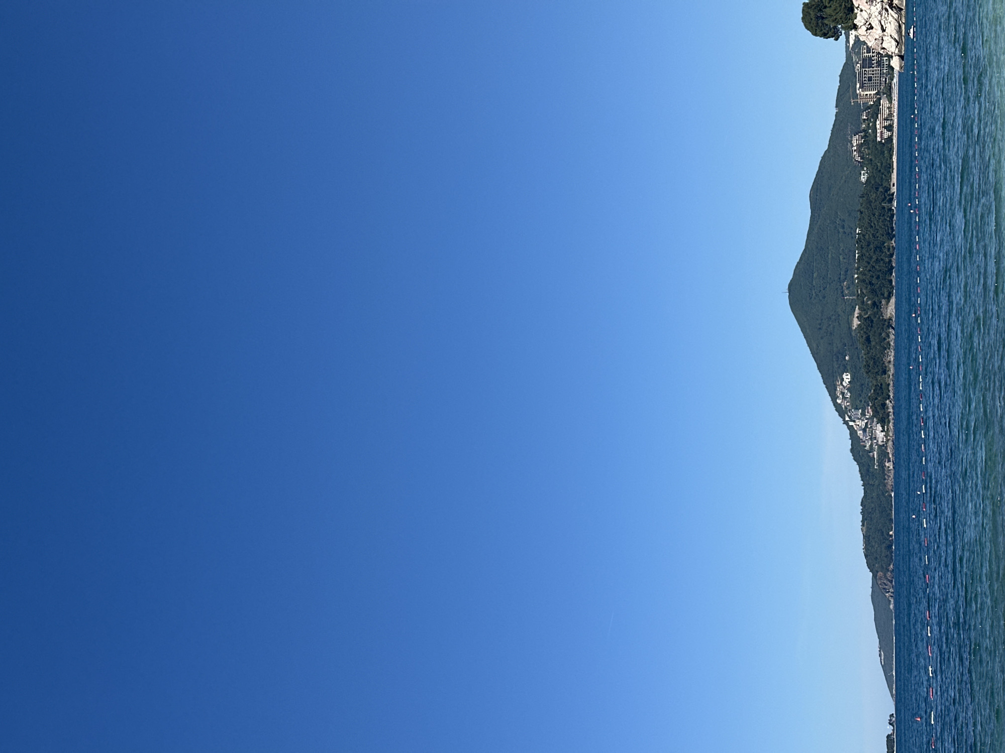 The image shows a view of Kamenovo, Montenegro. The sky is clear and blue. On the right side of the frame, there's a portion of the coastline with a tree on top of the rocks. A mountain range covered in vegetation descends to the coastline, where buildings are visible. The water appears to be calm, with a line of buoys visible on the surface. The landscape is bathed in sunlight.