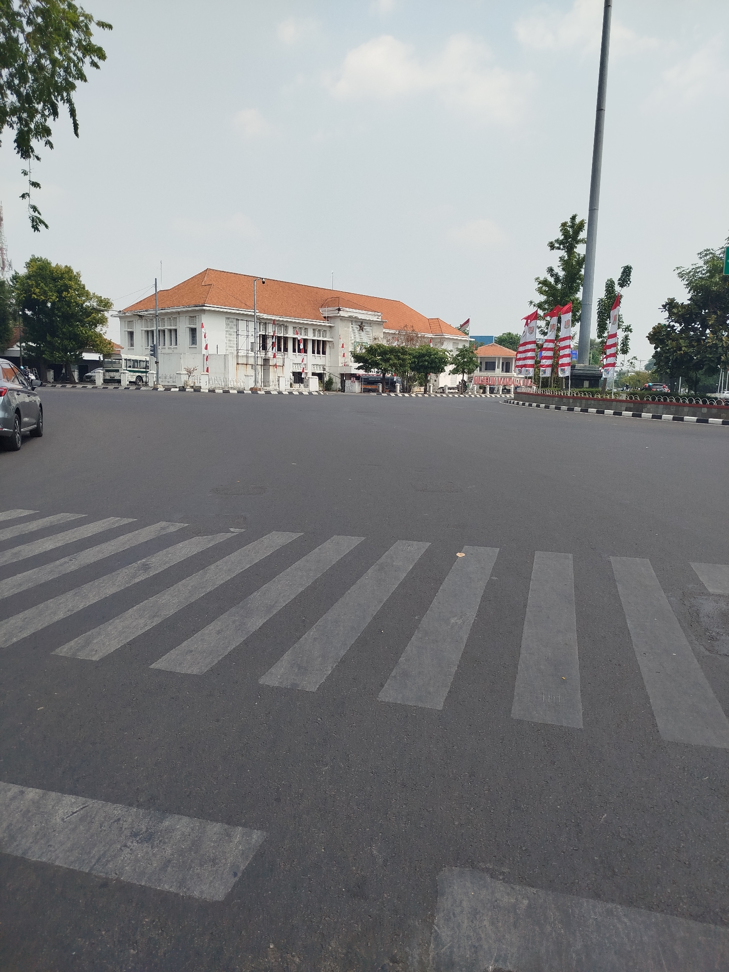 The image shows a wide asphalt road with a pedestrian crossing in the foreground. In the background, a white building with an orange-tiled roof is visible, adorned with Indonesian flags. The building appears to be a public institution, possibly a museum or government office, given its architectural style and the flags. A tall flagpole stands prominently on the right side of the frame, also decorated with flags. Trees line the street, and a cloudy sky completes the scene. The environment appears to be an urban setting in Semarang, Indonesia, possibly the area around the Lawang Sewu building, which is a well-known landmark with similar architectural features and often decorated with Indonesian flags.