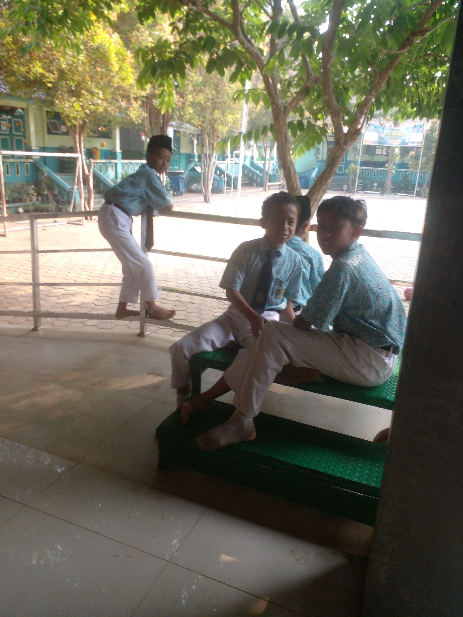 Four young boys, likely students, are present in an outdoor school environment. Three boys are sitting on green tiered seating, while one boy stands leaning against a railing. The boy standing is wearing a dark blue patterned shirt, white pants, and a peci (a cap common in Indonesia). The three sitting boys are wearing similar light blue patterned shirts and white pants. One of the sitting boys has a dark tie visible. The background shows a school building with a light blue facade and white trim, along with trees and fencing. The ground appears to be paved with light-colored tiles. The overall scene suggests a casual moment during the school day, possibly during a break or assembly, in Banyuputih, Indonesia.