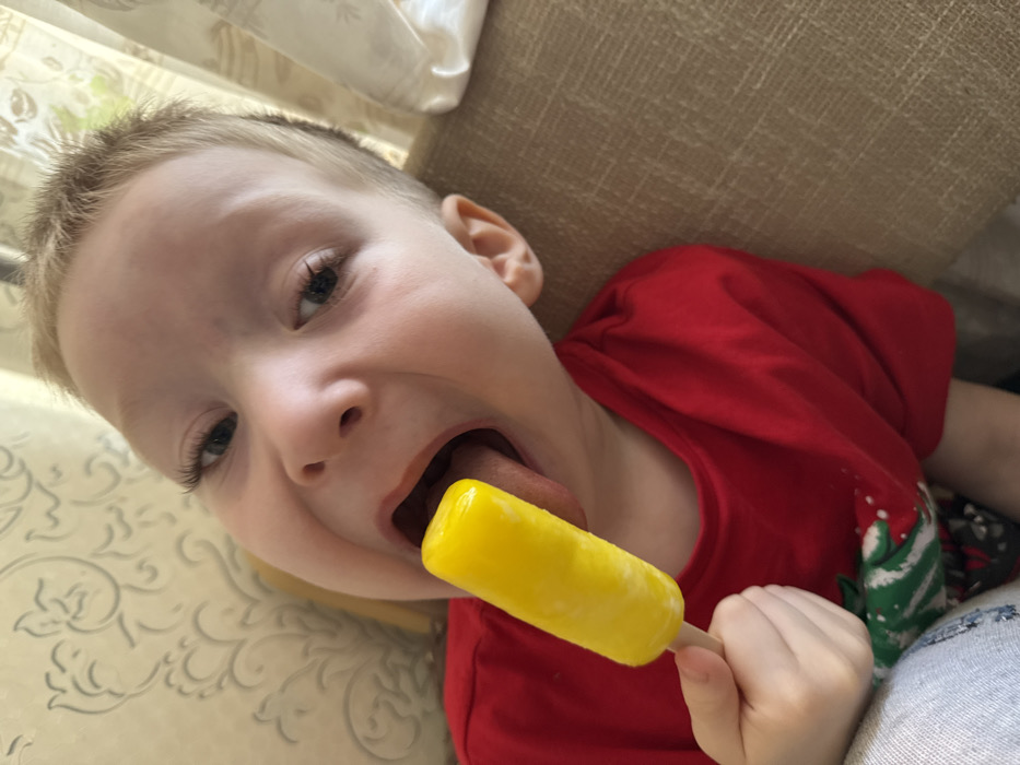 A close-up, slightly tilted image depicts a young child consuming a yellow popsicle indoors. The child, appearing to be a toddler or preschooler, has light-colored hair, fair skin, prominent eyelashes, and light-colored eyes. The child's mouth is open wide, with the tongue extended to make contact with the bright yellow, cylindrical ice pop, which is affixed to a white wooden stick. The child's right hand grasps the stick. The child is wearing a red short-sleeved t-shirt, with a partial glimpse of a green and white pattern visible on the lower left portion of the shirt. Grey fabric is visible in the bottom left foreground, possibly clothing. The background includes a light-colored wall with a repeating grey floral or vine pattern on the left side. To the right, a textured light brown or beige upholstered surface, consistent with a couch, is visible, along with a light-colored, possibly sheer, curtain. The scene takes place within a private residential interior; specific identifying markers for the city of Chaikovsky, Russia, are not visible within the image.