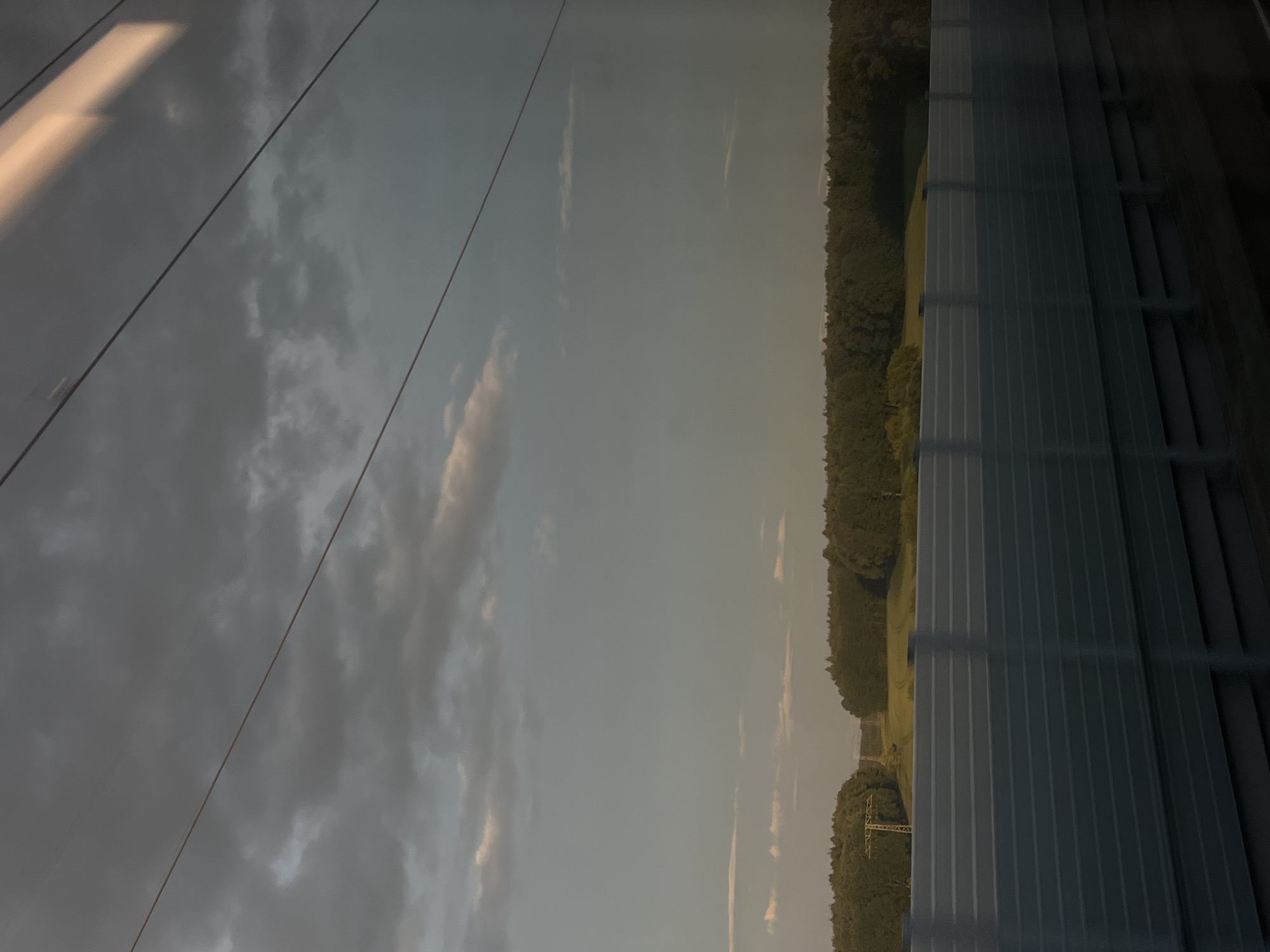 The image depicts a view through a window, likely from a moving vehicle such as a train, looking out over a landscape. In the foreground, a light-colored, horizontally paneled barrier extends across the width of the frame, obscuring the lower portion of the scene. Above this barrier, a dense line of dark green trees forms the horizon. Beyond the trees, a field with patches of yellow and green is visible, interspersed with more trees. In the far distance, there are hints of structures, possibly power lines or small buildings, along with distant hills or a lighter sky. The sky itself is a mix of muted grays and blues, with scattered clouds, some darker and more ominous, others lighter and wispy. A bright, elongated streak of light, possibly a reflection or sunlight filtering through, is visible in the upper right portion of the sky. Several thin, dark lines, likely overhead wires, crisscross the sky from left to right. The overall impression is of a rural or semi-rural landscape under a cloudy sky. The provided context of Ilmenau, Germany, suggests this is a view from a location within or near that city.