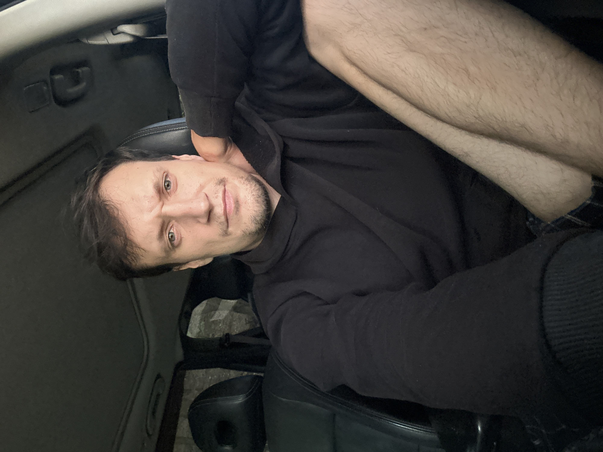A man with brown hair, stubble, and light blue eyes is sitting in the driver's seat of a car. He is wearing a black hoodie and plaid shorts. His left arm is resting on the headrest behind him, and his right leg is bent and resting on his left. The car's interior is dark, with leather seats and a sunroof visible. The background outside the car appears to be a dimly lit, outdoor environment, possibly at night. The man's facial expression is neutral. There is no indication of the specific location within the city of Makiv, Ukraine, other than it is inside a vehicle.