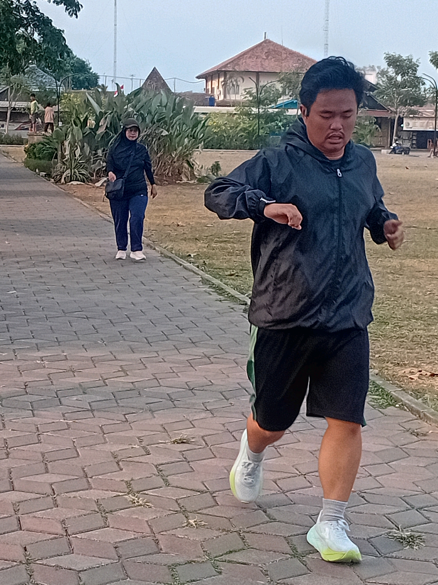 In Gebog, Indonesia, a man is captured mid-stride, jogging on a paved walkway. He is wearing a dark hooded jacket, black shorts with a green stripe, light-colored socks, and white running shoes with a neon yellow accent on the sole. His gaze is directed forward, and he appears to be wearing earbuds or headphones. Behind him, a woman dressed in a black hijab and dark clothing, also wearing white sneakers, walks on the same path. The walkway is constructed of interlocking rectangular pavers. The surrounding environment features dry grass, scattered green plants, trees, and in the background, a building with a reddish-brown roof and other structures. The sky is overcast.