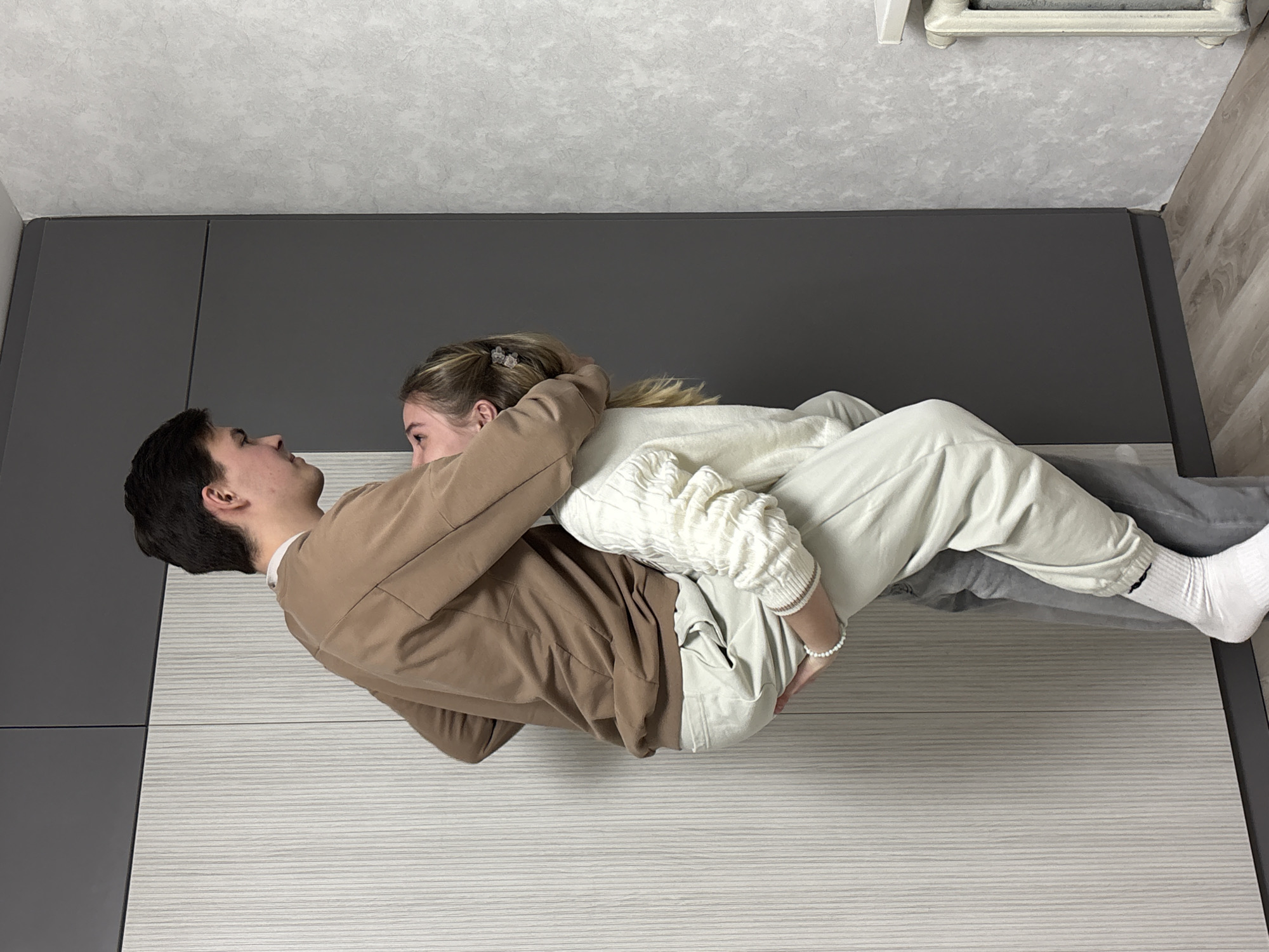 Two young individuals, a male and a female, are positioned intimately on a light grey striped surface, likely a bed or large mat, in an indoor setting in Chaikovsky, Russia. The male, with dark hair and wearing a brown long-sleeved top and grey trousers, lies on his back, looking upwards towards the female. The female, with blonde hair tied back and adorned with a white beaded bracelet on her left wrist, is situated on top of him, wearing a cream-colored cable knit sweater and light-colored trousers. She is looking down at the male, their eyes meeting in a tender gaze. Their arms are wrapped around each other, suggesting an embrace.
The background features a dark grey wall directly behind the couple, and a light grey textured ceiling above them. To the upper right, a white radiator is partially visible, next to a light wooden floor. The overall scene conveys affection and intimacy within a modern interior environment. There is no visible text, and the time of day or weather cannot be determined from the image.