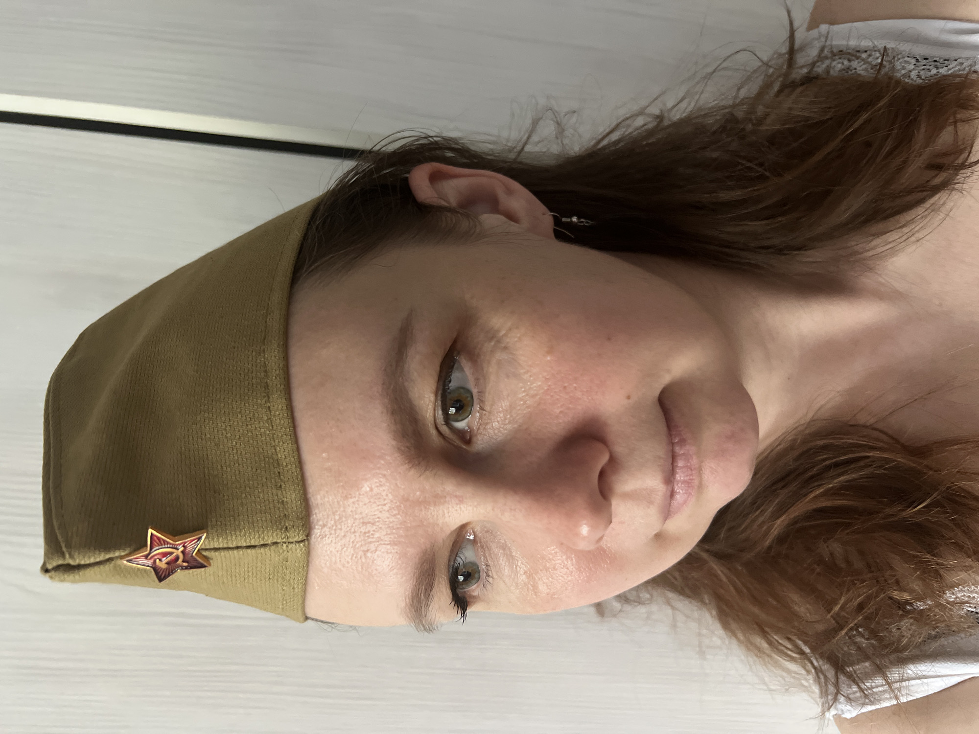 A woman is wearing a Soviet-era military cap, complete with a red star badge bearing a hammer and sickle emblem, on a white wooden surface. The background includes a strip of the same white wooden surface and what appears to be a white wall with a black line dividing it. The photo was not necessarily taken in Chaikovsky, Russia.