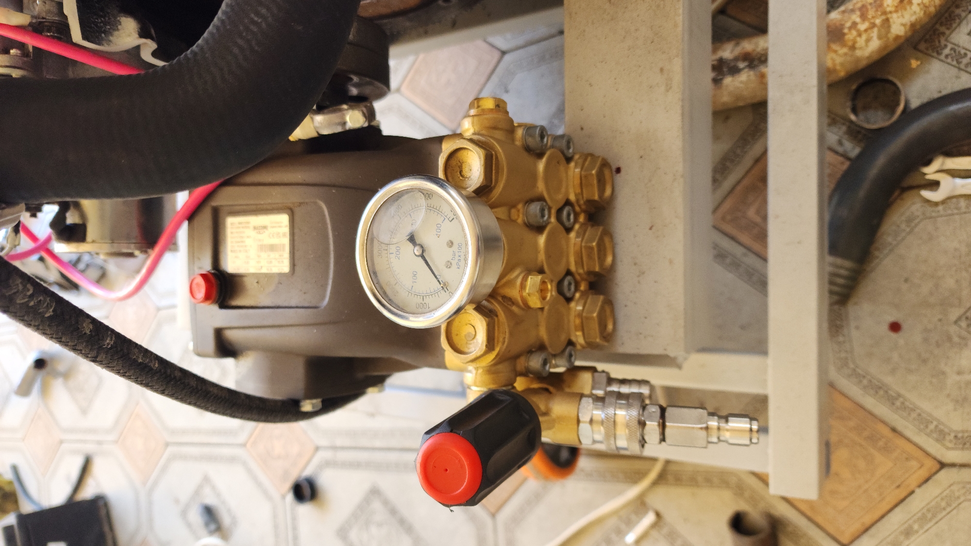 The image shows a close-up view of a pressure washer pump and its components. The pump itself is a gold-colored metal unit with multiple hexagonal fittings and bolts. A pressure gauge with a white face and black markings is attached to the pump, displaying a reading around 100 bar. Below the gauge, a black knob with a red cap is visible, likely a pressure adjustment or control.
Connected to the pump are thick black hoses, one of which is partially visible at the top left of the frame, and another that extends down the left side. Red wires are also seen, suggesting an electrical connection. A label with text is affixed to the main body of the pump.
The background suggests an outdoor setting, possibly a workshop or a yard, with patterned floor tiles visible. There are hints of other machinery or equipment in the background, including rusty pipes and a dark curved hose. The lighting indicates it is daytime. The scene appears to be a still shot of equipment, with no activity or people present.
The text on the label is partially legible, with "BAR" and "kPa x 100" visible on the gauge, indicating pressure units. The label on the pump contains technical specifications or identification details, but the text is too small and blurry to be clearly read. The overall impression is a detailed look at the functional parts of a pressure washing system.