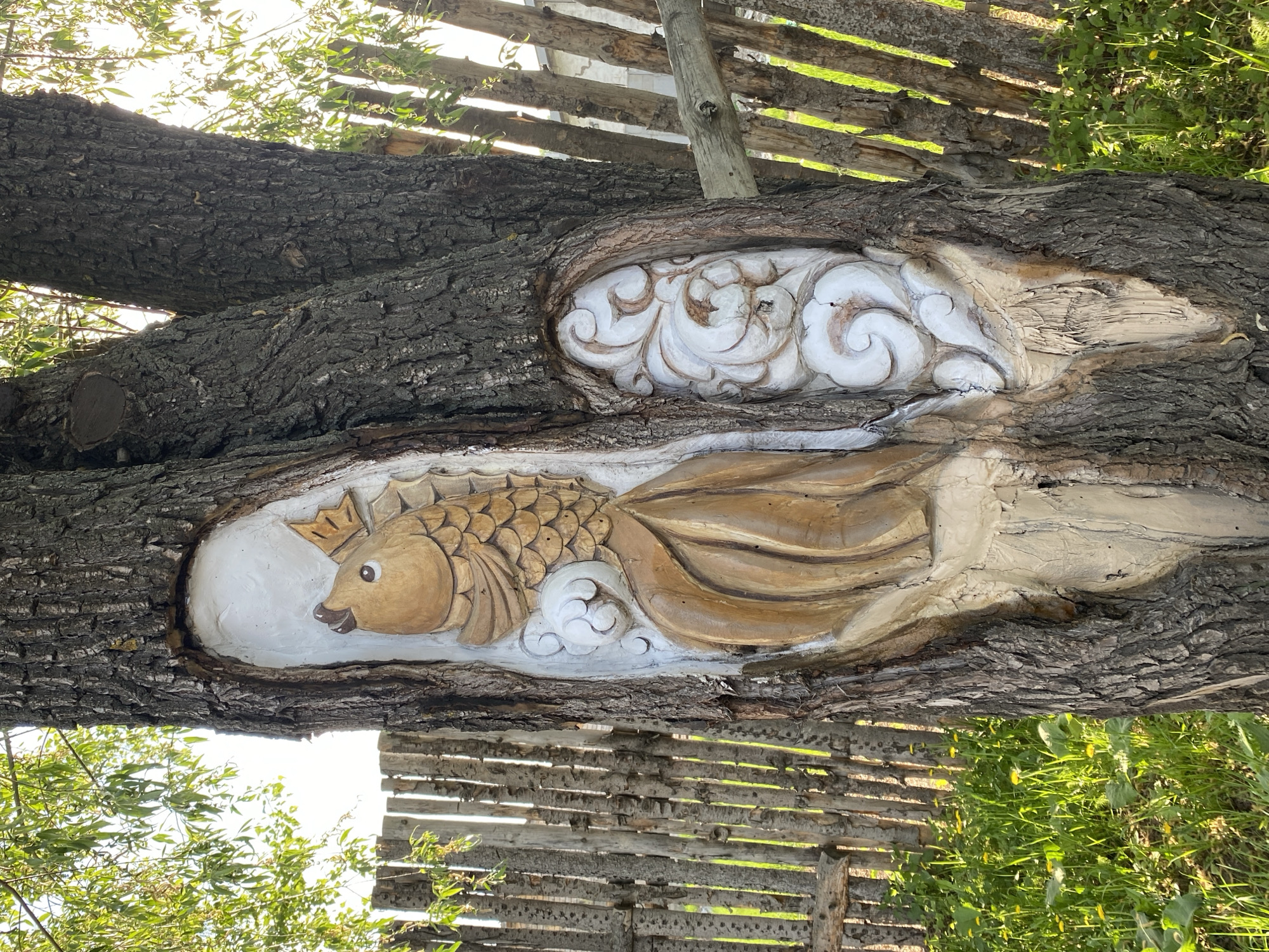 A horizontally-oriented, medium-shot photograph shows a carved tree trunk with two oval-shaped carvings. The upper carving features white, stylized floral or cloud-like designs. The lower carving depicts a brown, carved fish with scales and fins, surrounded by white swirls. The tree trunk is brown with rough bark. In the background, there are glimpses of a wooden fence, greenery, and sky. The photograph is taken in the city of Chaikovsky, Russia.