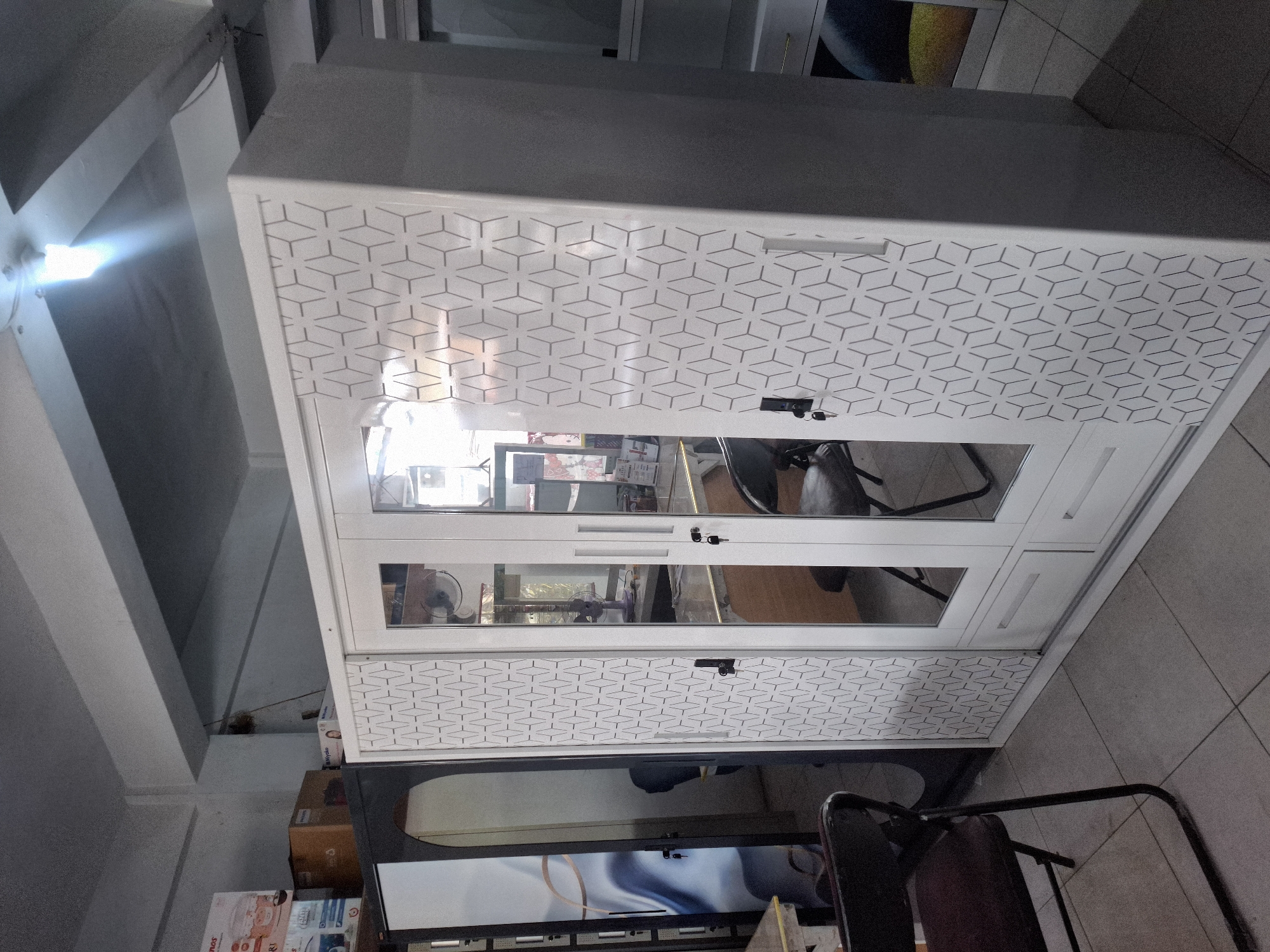 The image shows the interior of a store, likely a furniture or appliance shop, in Banjarmadu, Indonesia. In the foreground, a large white wardrobe with mirrored doors and a geometric pattern is prominently displayed. To the left of the wardrobe, a dark metal folding chair with a maroon seat is visible. The floor is tiled with light gray square tiles. In the background, other furniture items, boxes, and a purple fan are partially visible. The lighting suggests overhead fluorescent lights. The overall impression is a retail environment with merchandise on display.