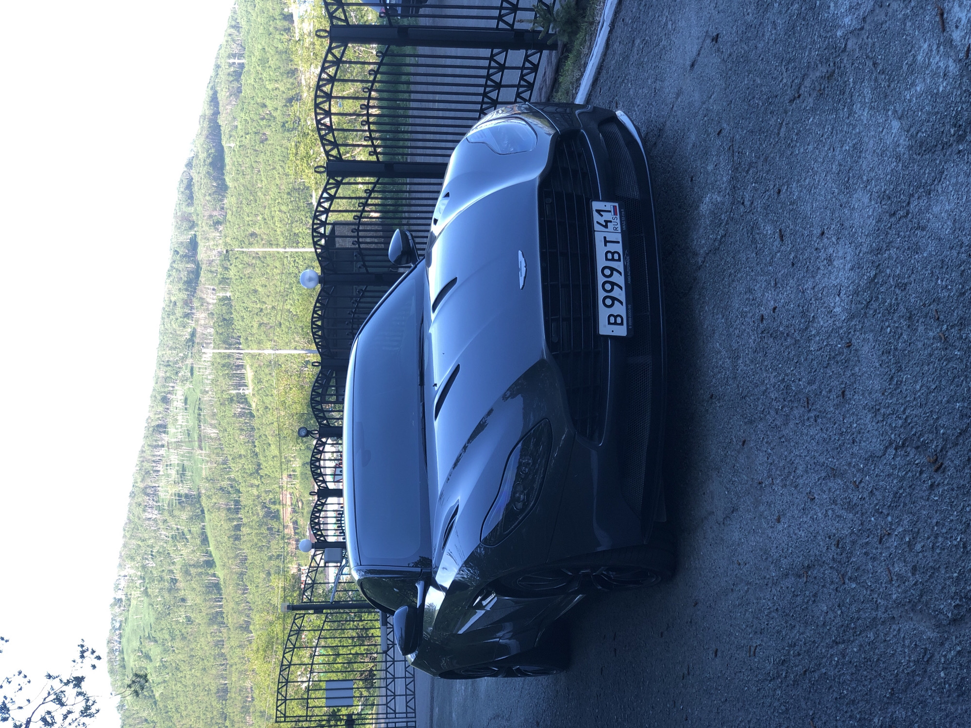 The image depicts a dark gray Aston Martin parked on an asphalt driveway in front of a black wrought-iron gate. Behind the gate, a lush green hillside covered in trees rises steeply. The sky is bright and overcast. A license plate on the Aston Martin reads "B 999 BT 41 RUS". The scene is likely located in Petropavlovsk-Kamchatsky, Russia, given the license plate format and the mountainous, verdant landscape which is characteristic of the region. There are no people visible in the image. The car is the primary subject, with the gate and the natural environment forming the backdrop.