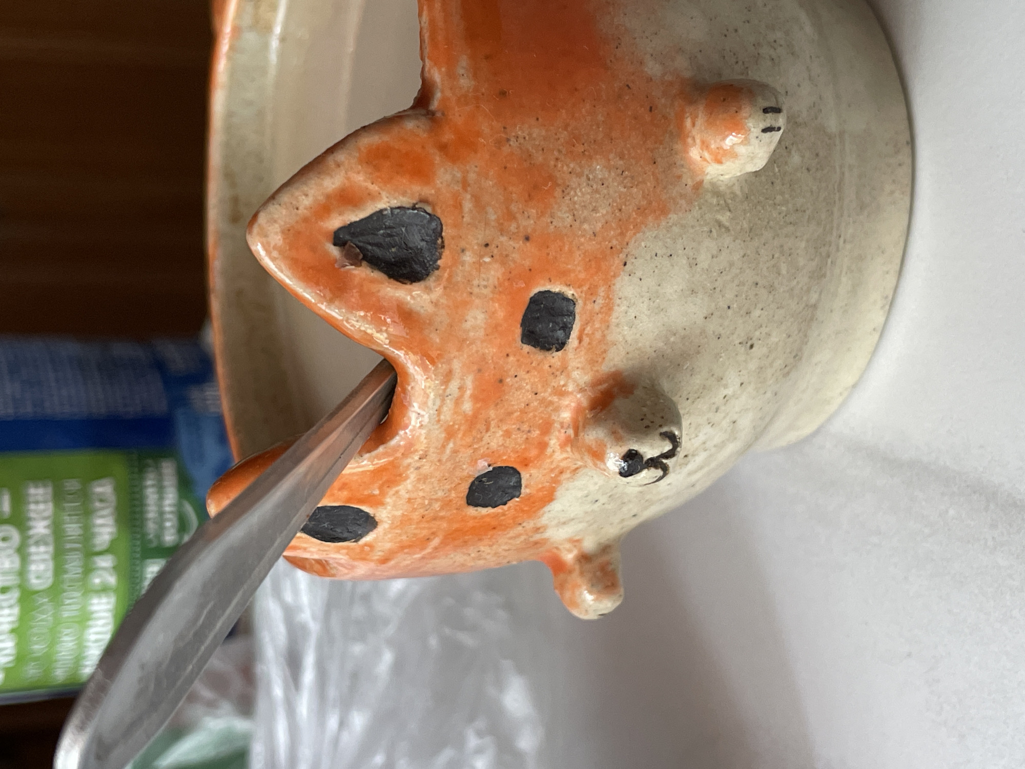 A close-up shot shows a handmade ceramic bowl in the shape of a fox, with an orange and white speckled glaze. The bowl is tilted at an angle. A silver spoon is inserted into the fox's mouth. The background is blurry, but includes a blue and green product package with Russian text and a crumpled clear plastic bag. The surface beneath the bowl appears to be a white countertop. There is no indication of the location being in the city of Zheleznogorsk, Russia. No people are visible.