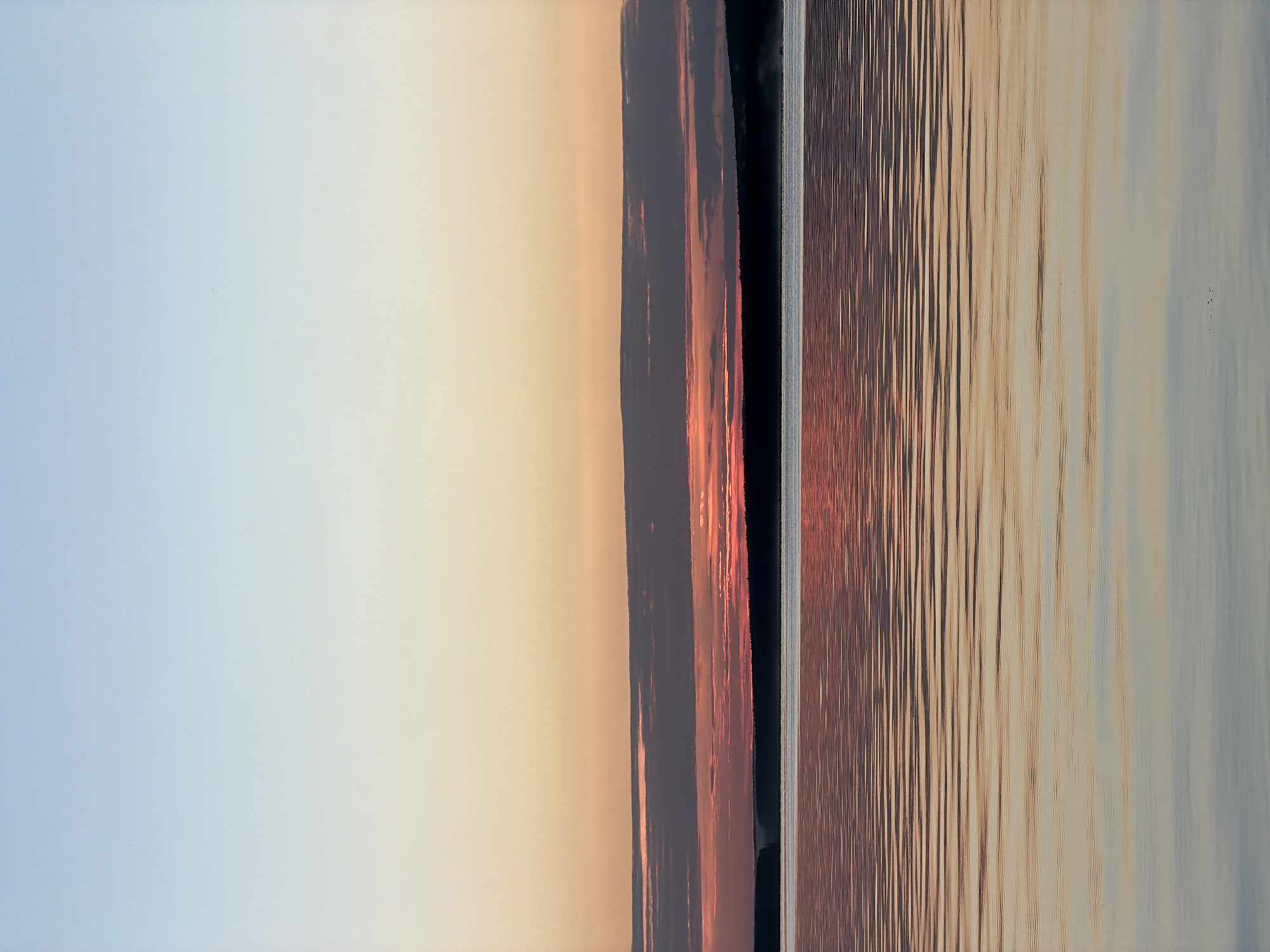 The image depicts a serene natural landscape at twilight, likely captured during sunset over a body of water. The sky transitions from a pale blue at the top to soft shades of orange and pink near the horizon, with a band of clouds tinged with red. Below the sky, a dark, silhouetted treeline runs horizontally across the frame, marking the edge of the land. The foreground is dominated by a calm body of water, reflecting the colors of the sky and clouds in gentle ripples. The water's surface exhibits a gradient of colors, from warmer hues near the horizon to cooler blues and grays in the lower portion of the image. There are no visible people, man-made structures, or specific landmarks that would pinpoint a location within Chaikovsky, Russia. The scene is purely natural and atmospheric.