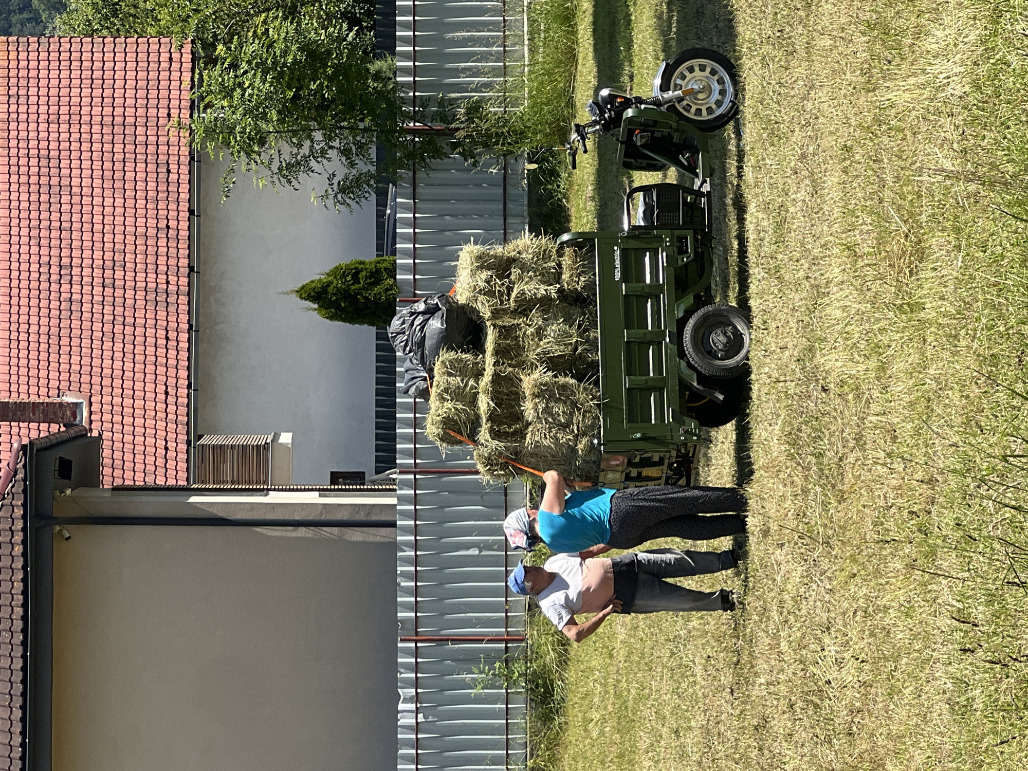 In an outdoor scene, two individuals, a man and a woman, stand near a green three-wheeled utility vehicle loaded with hay bales and a black plastic bag. The man, wearing a light-colored t-shirt, dark pants, and a blue cap, stands with his hands on his hips. The woman, in a blue top and patterned pants, faces away from the camera and appears to be holding onto a hay bale. Behind them, a metal fence runs horizontally across the image. In the background, a building with a reddish-brown tiled roof is visible, along with some trees and greenery. The ground is covered in dry grass.