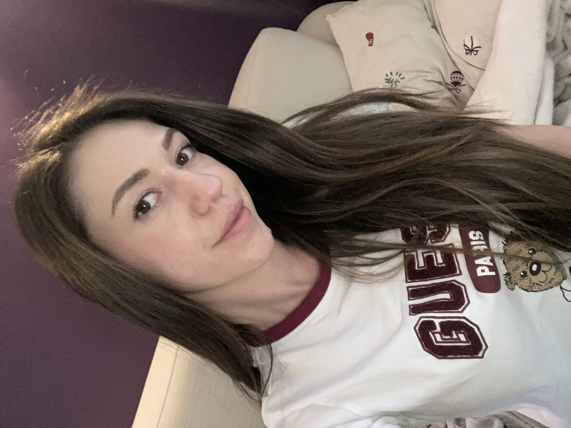A young woman with long, dark brown hair is captured in a close-up, slightly angled selfie. She has fair skin, dark eyes, and a subtle, soft smile, looking directly at the camera. Her hair is spread out around her head, with some strands appearing slightly disheveled near her forehead. She is wearing a white ringer t-shirt with a maroon collar.
The t-shirt features text and a graphic design; visible words include "GUES" (likely part of "GUESS") in large, maroon block letters, and "PARIS" written vertically in maroon next to it. A small, stylized brown bear's head graphic is also visible next to the "PARIS" text.
She appears to be reclining or lying down on a light-colored surface, possibly a sofa or bed. In the background, behind her head, there is a solid dark purple wall. Adjacent to her hair, several light-colored cushions or pillows are visible, one of which has small, embroidered designs in maroon and green, resembling a hot air balloon, a gift, and a plant. The overall setting appears to be indoors, with soft lighting. The atmosphere suggests a relaxed and casual moment.