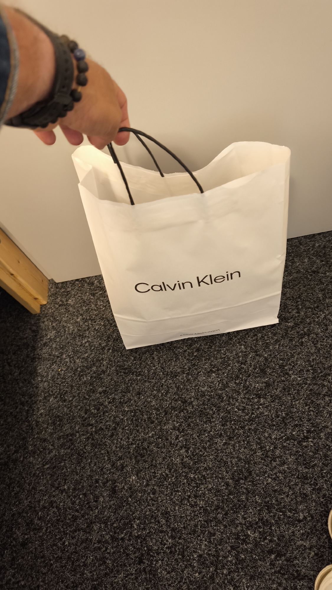 A medium-shot photograph shows a white Calvin Klein shopping bag being held by a hand. The bag has black handles and the Calvin Klein logo printed in black. The hand holding the bag has a dark-colored watch and a bracelet made of dark-colored beads. The bag is sitting on a gray carpet. A white wall is visible in the background, along with a portion of a wooden frame near the bottom left corner of the frame. The image was taken indoors. The location is Unknown, Unknown.